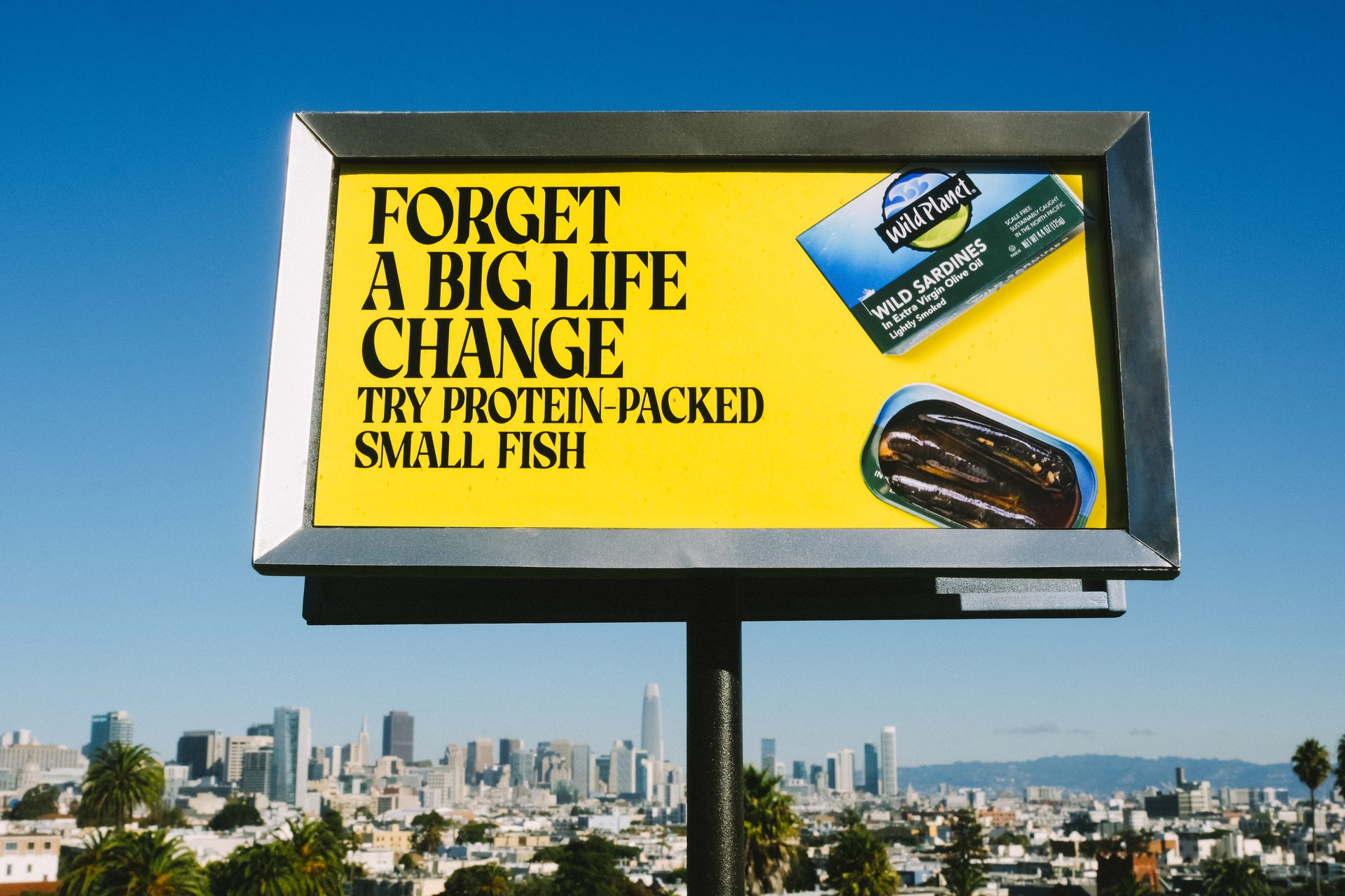 A bright yellow billboard displays an ad for Wild Planet sardines against a blue sky, overlooking a cityscape.