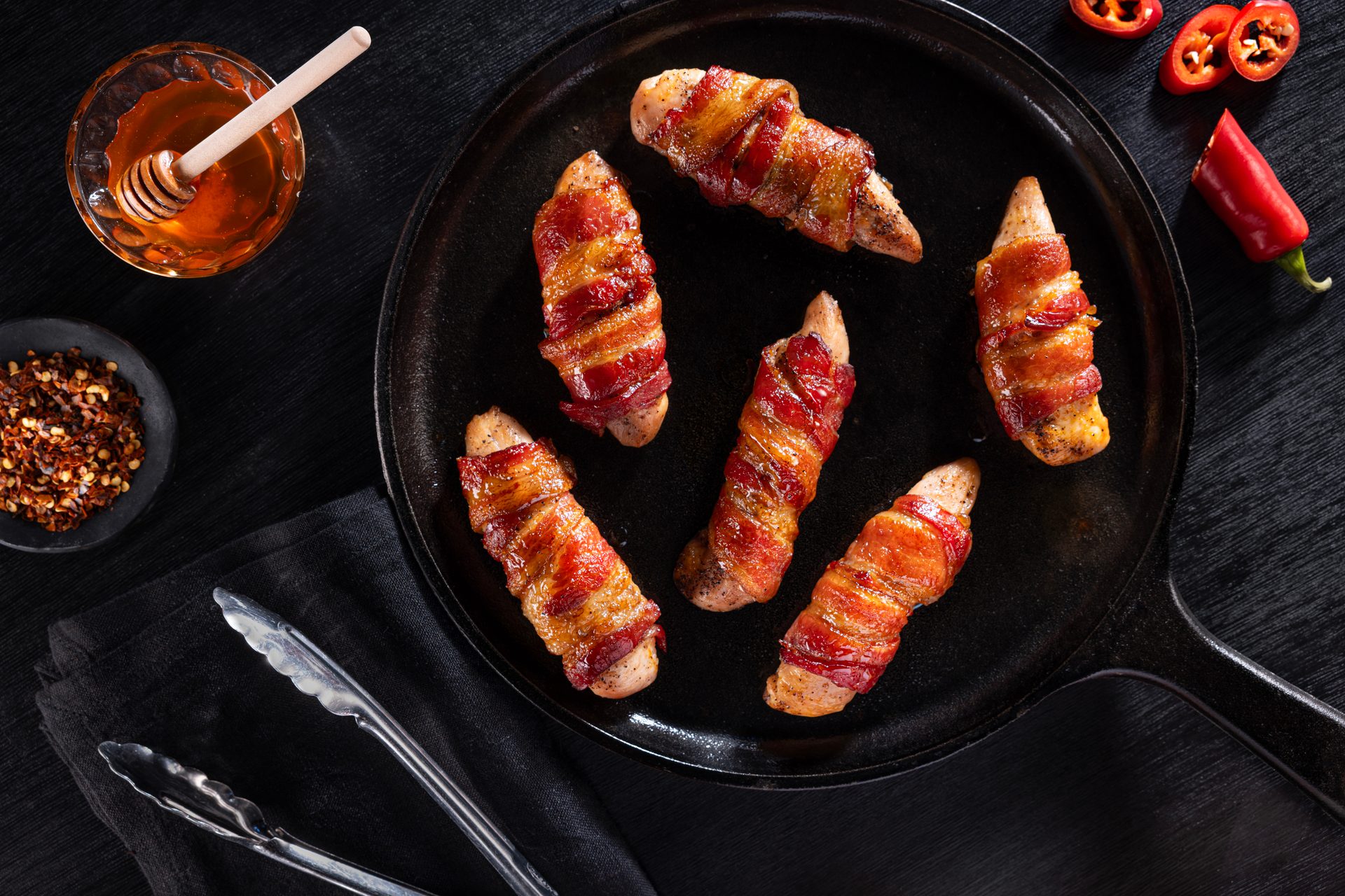 Overhead shot of crispy bacon-wrapped chicken in a skillet, with honey, chili flakes, and red chilies.