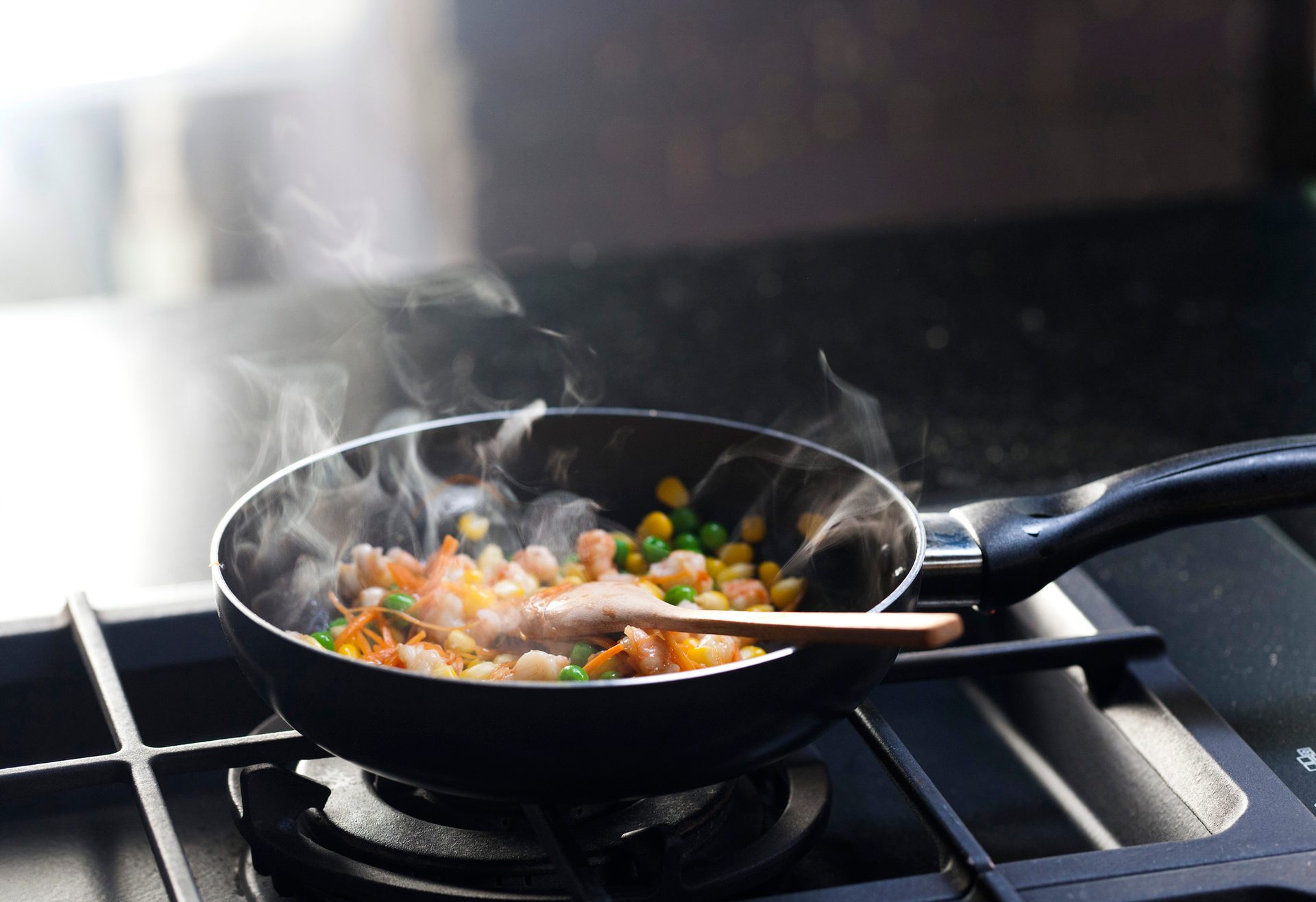 Frying pan on a gas stove, filled with steaming colorful vegetables and shrimp, stirred by a wooden spoon.
