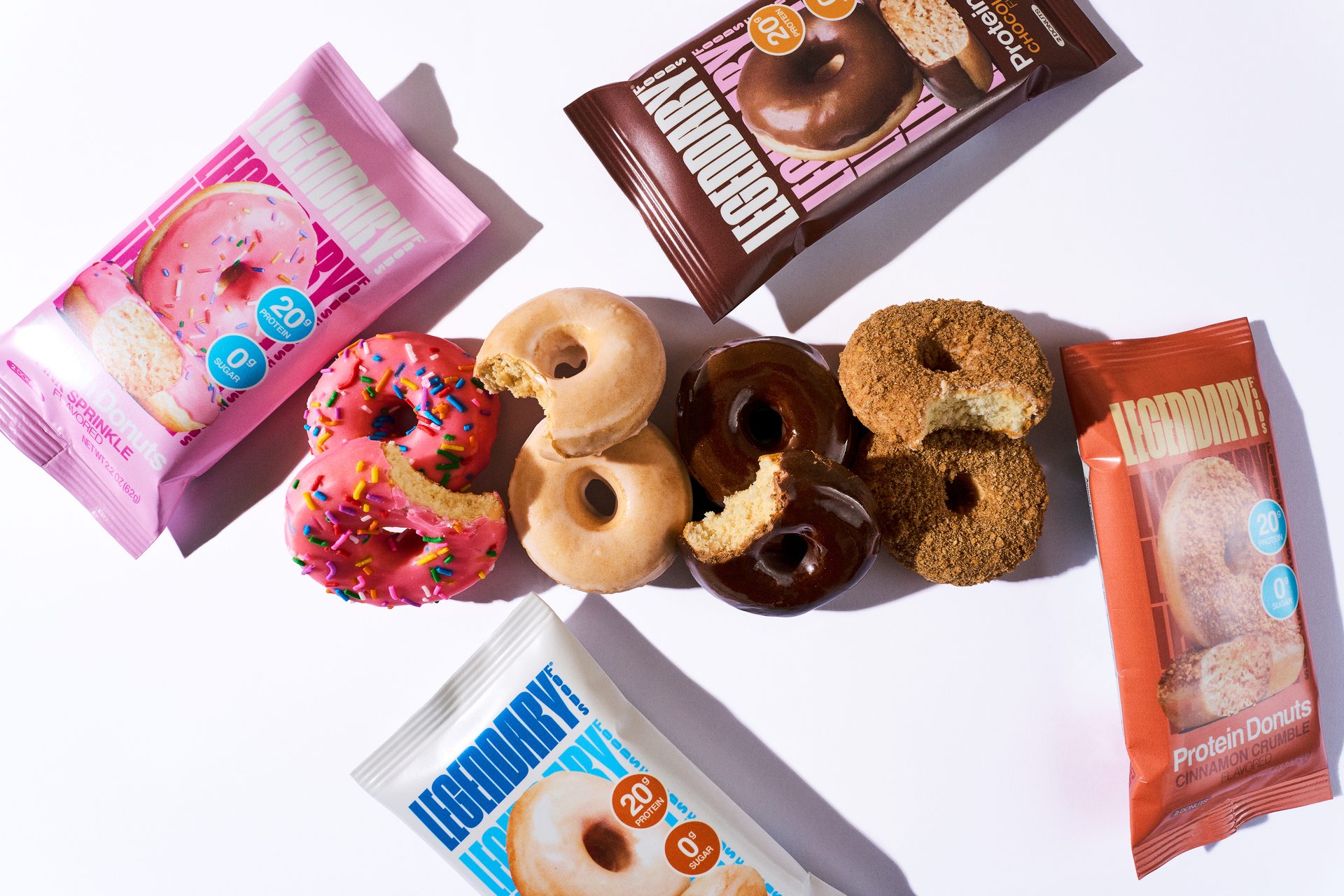 Various flavors of Legendary Foods protein donuts and their colorful packaging, some with bites taken out.
