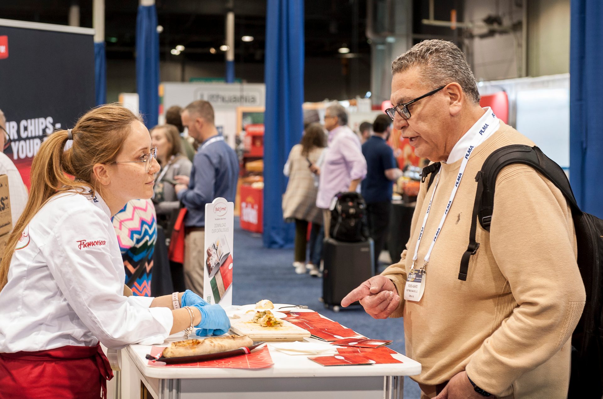 A woman in a chef's coat engages a man in a beige sweater at a busy trade show food booth.