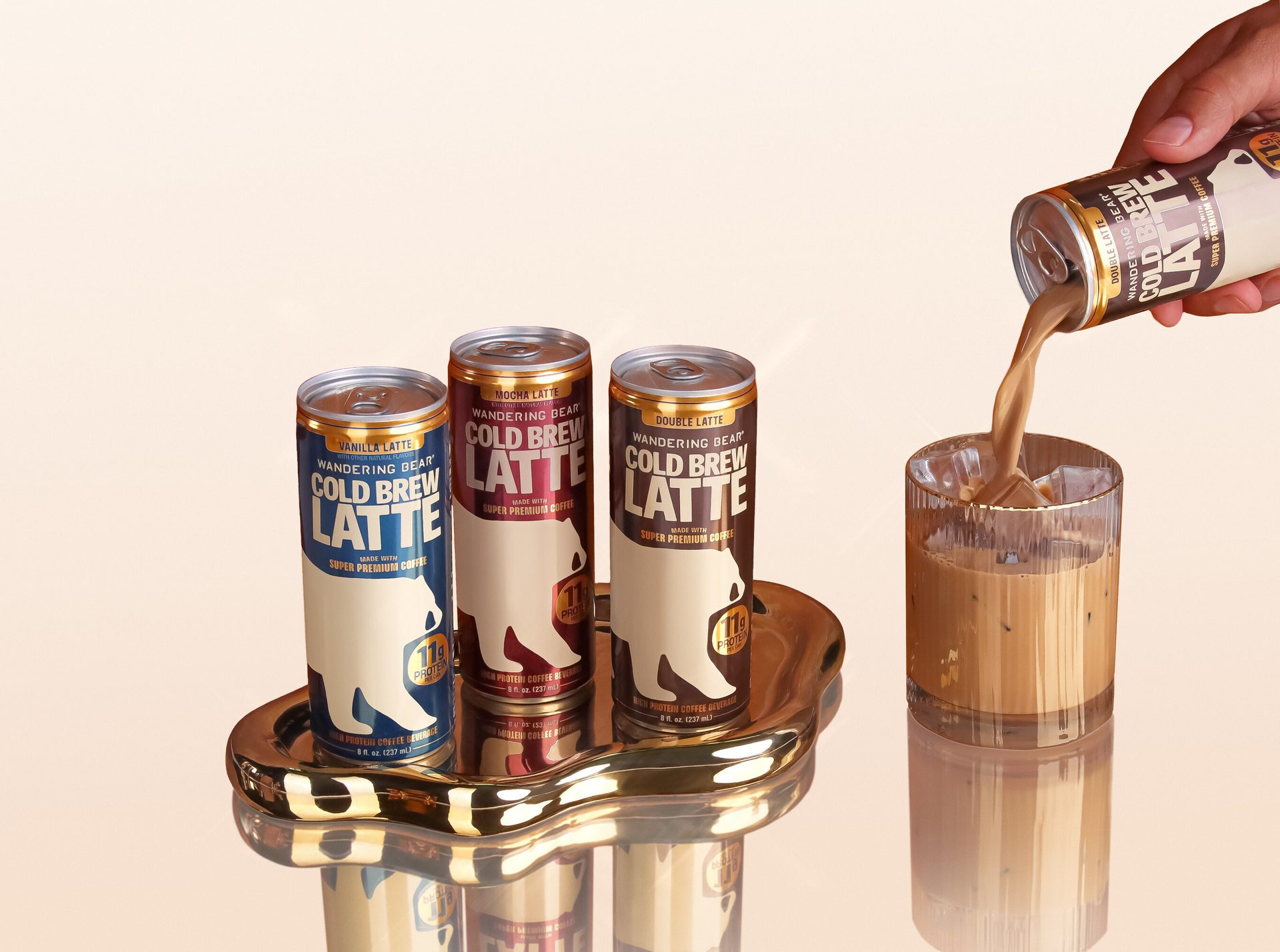 Three Wandering Bear cold brew latte cans on a gold tray, one being poured into an iced glass.