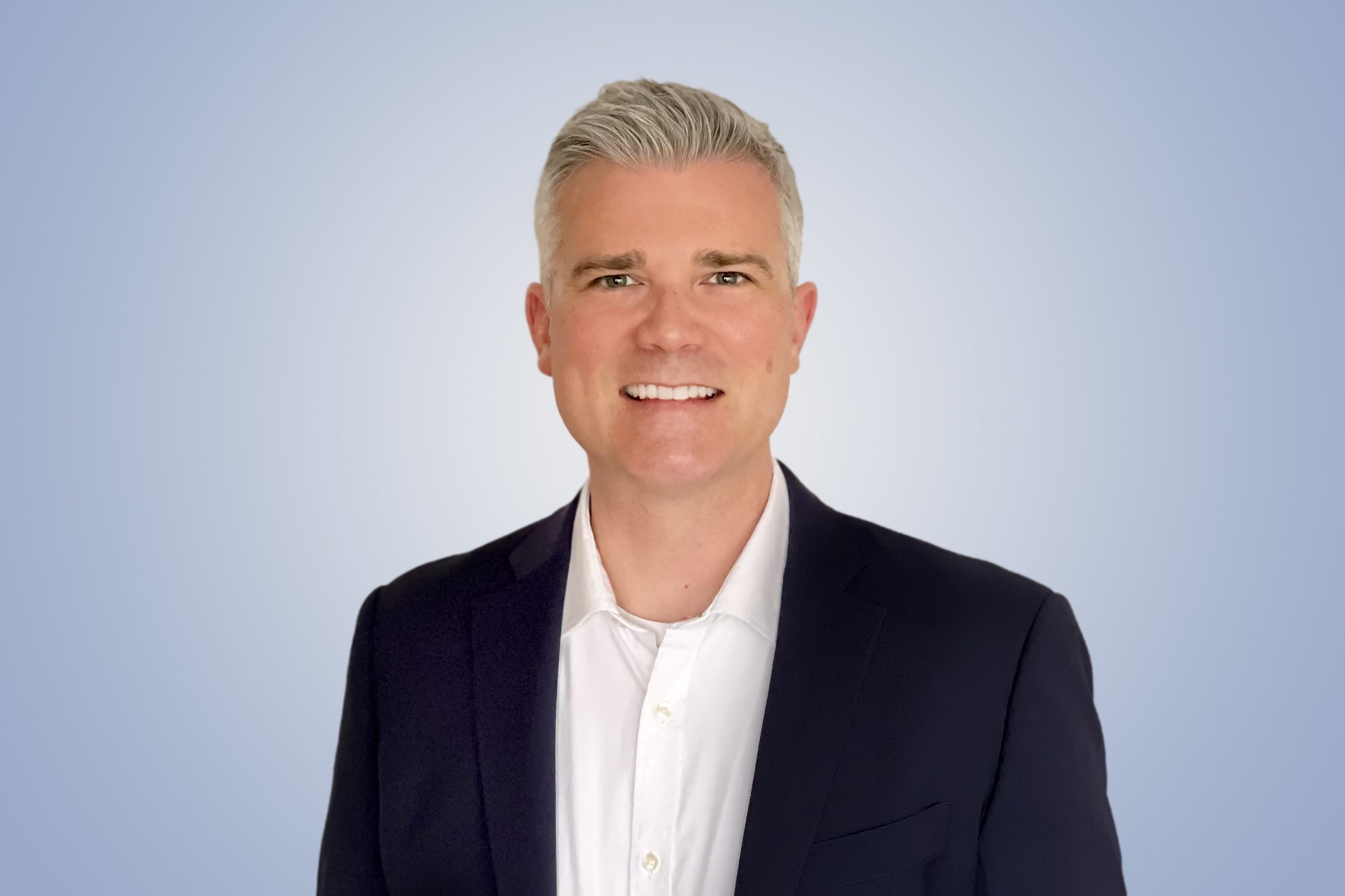 Smiling man with gray hair in dark suit, light blue background.