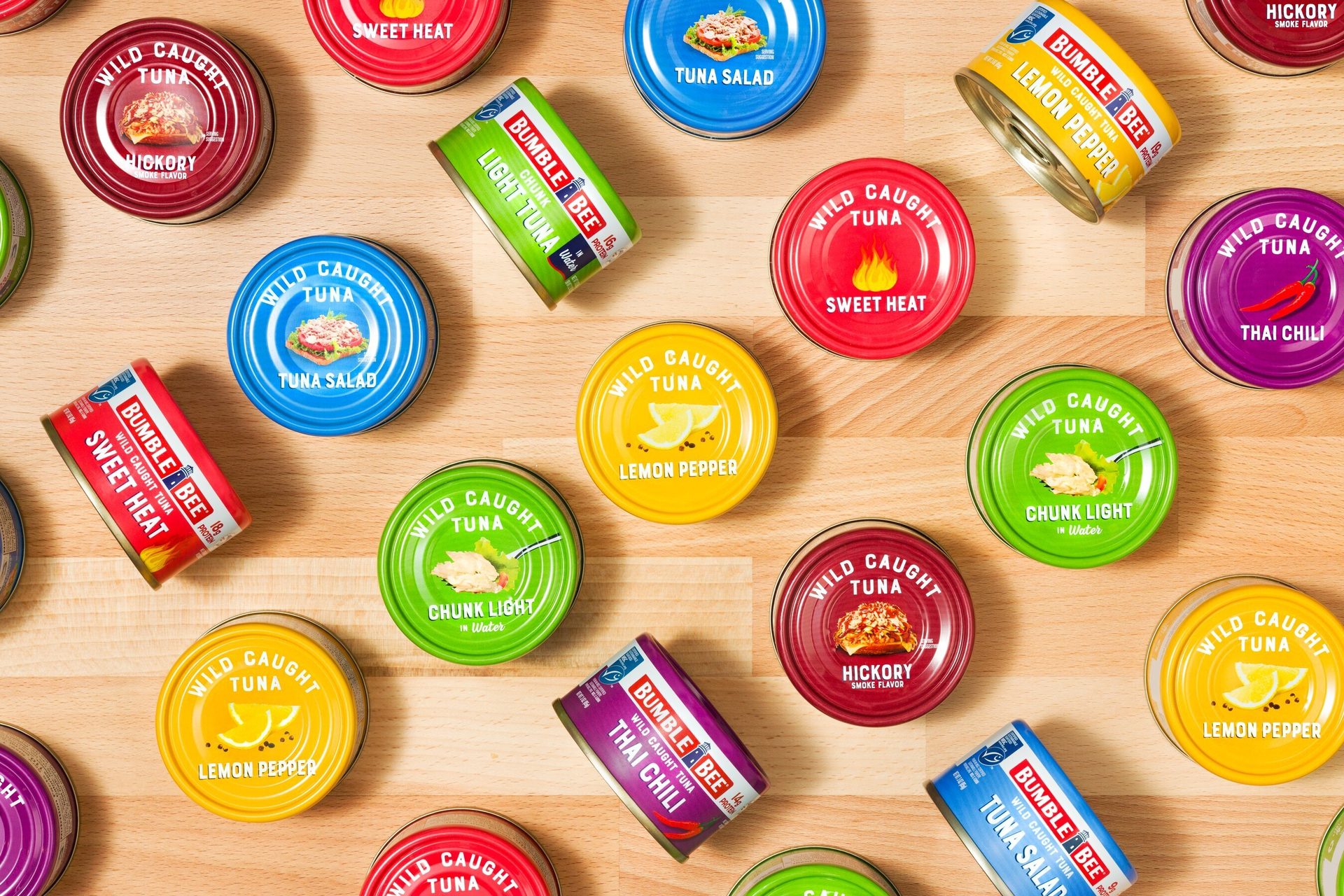 An overhead shot of numerous colorful Bumble Bee tuna cans in various flavors scattered across a wooden surface.