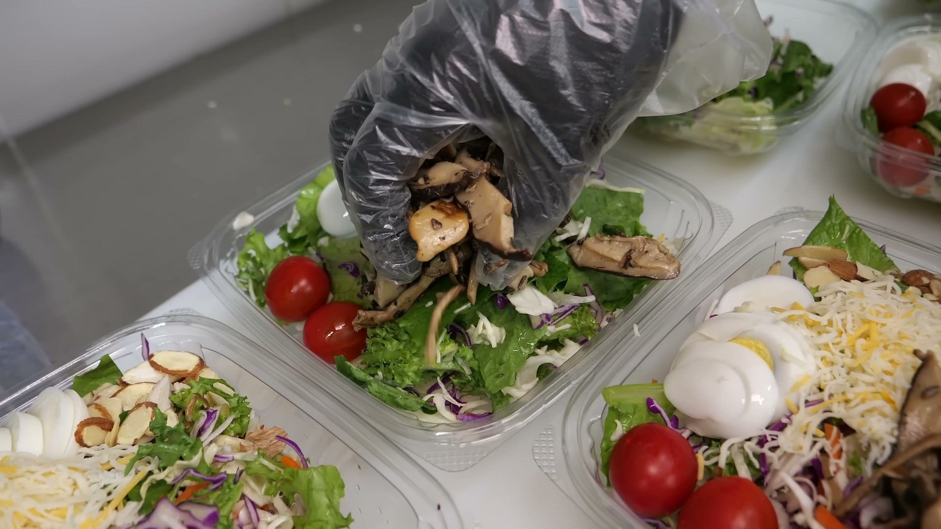 Hand in a clear glove adding sautéed mushrooms to a fresh green salad in a plastic container.