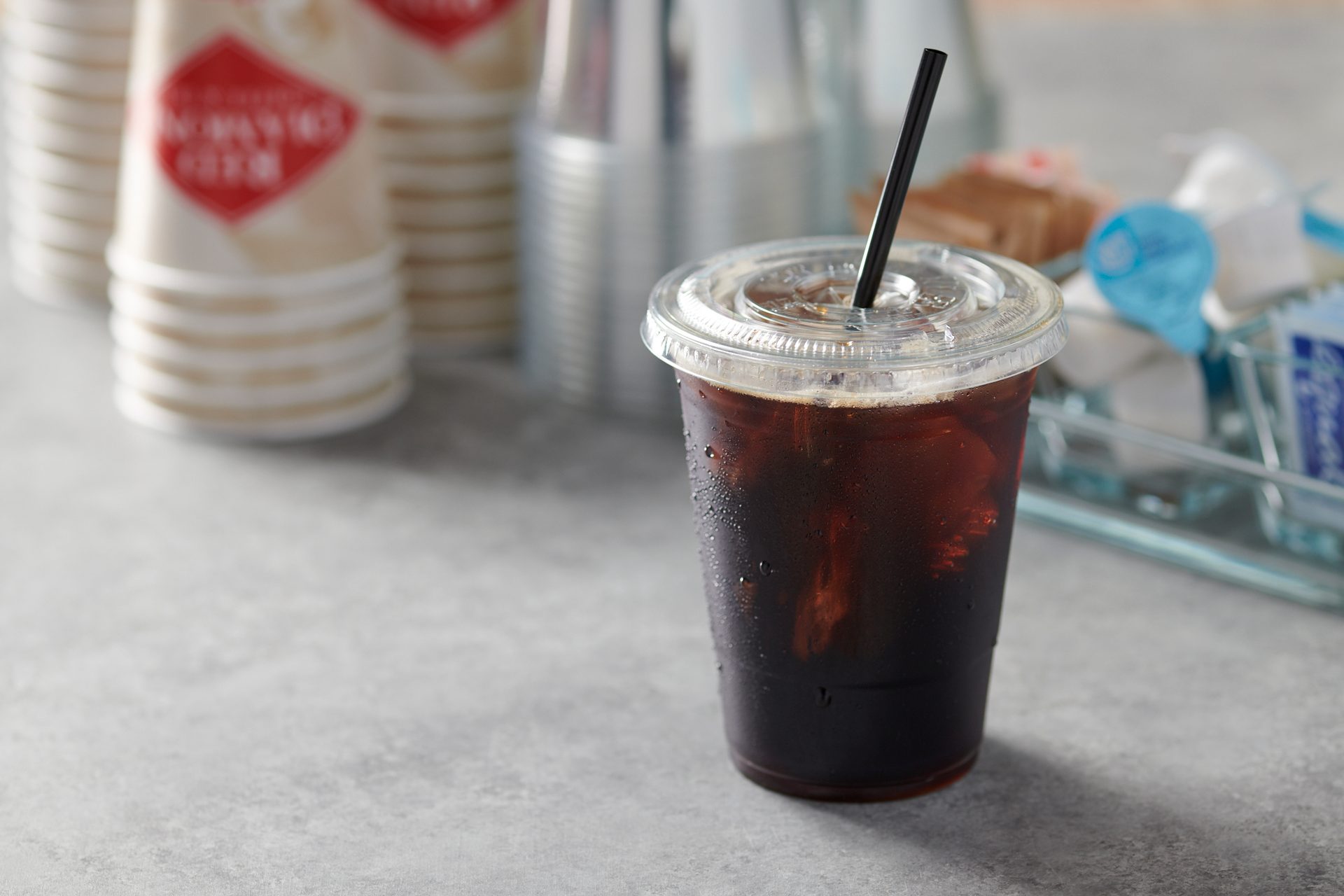Iced coffee with straw in clear cup, condensation, blurred coffee cups and condiments.