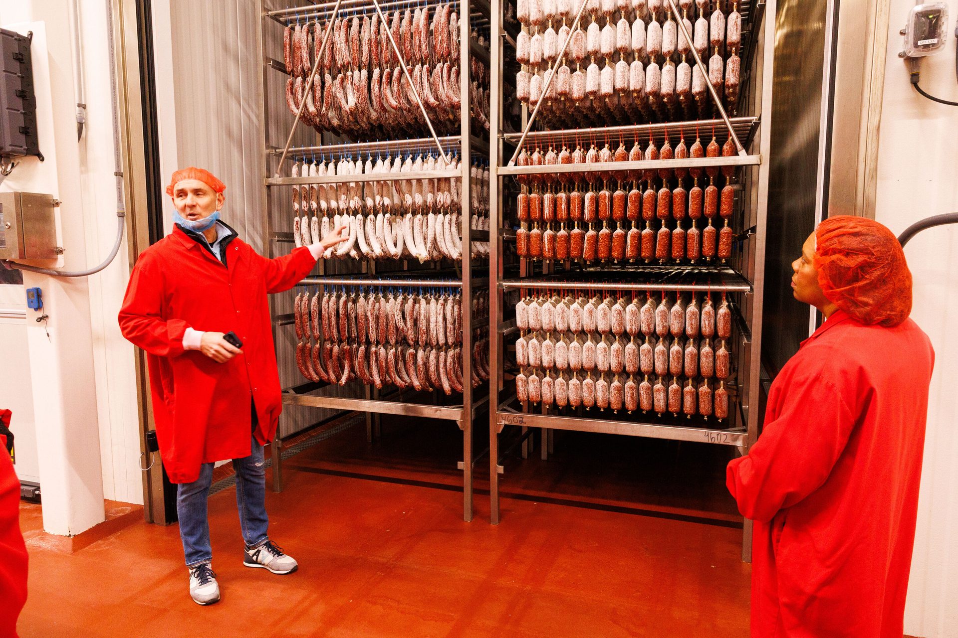 Two workers in red coats and hairnets inspect racks of drying sausages in a food processing plant.