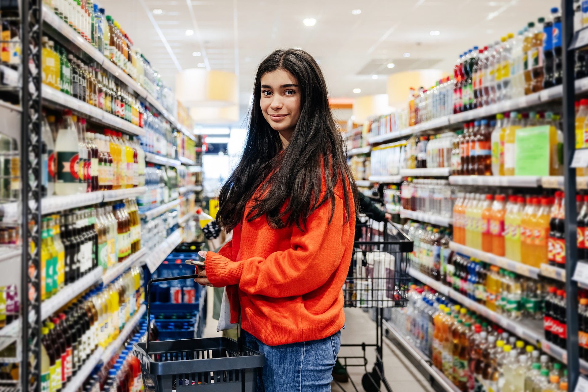 Smiling young woman in a red sweater holds a shopping basket in a grocery aisle full of drinks.
