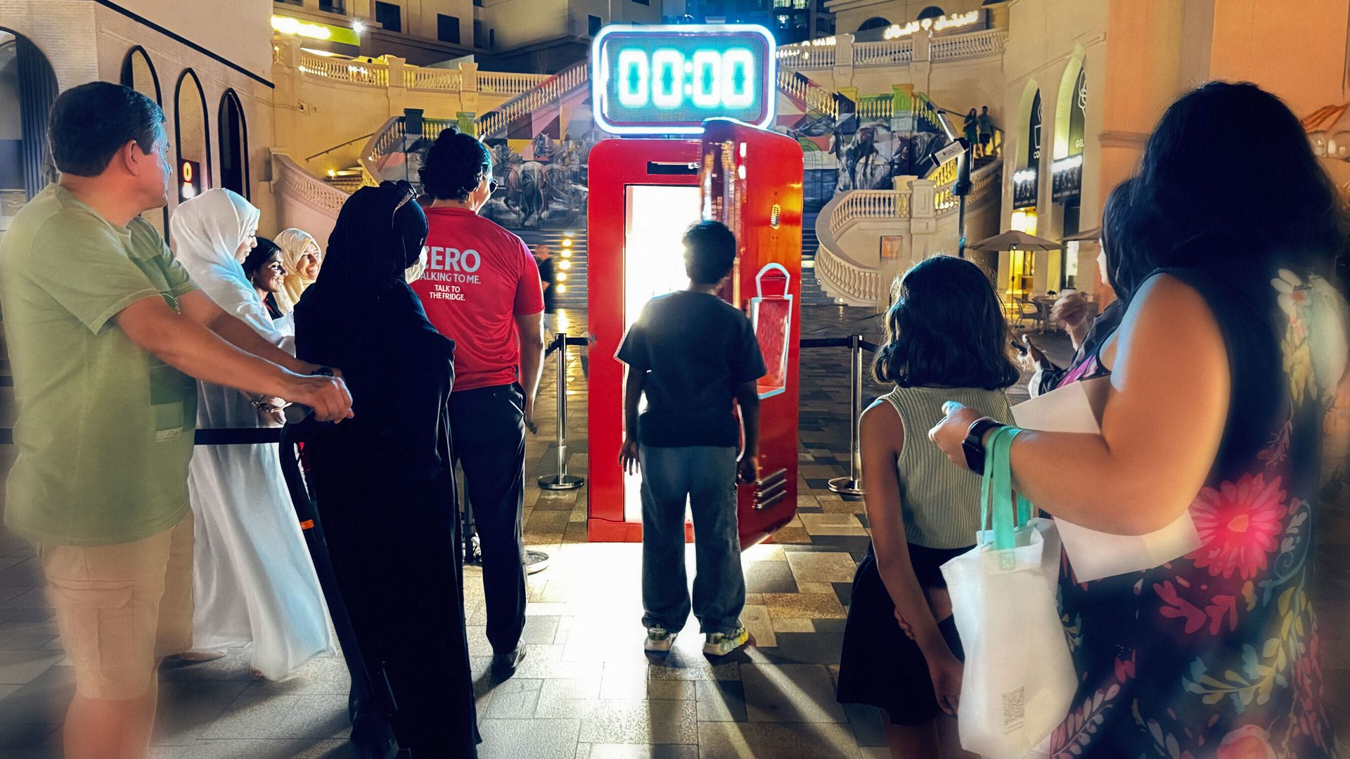 People gathered around a red interactive vending machine with a digital timer set to 00:00, in an outdoor evening setting.