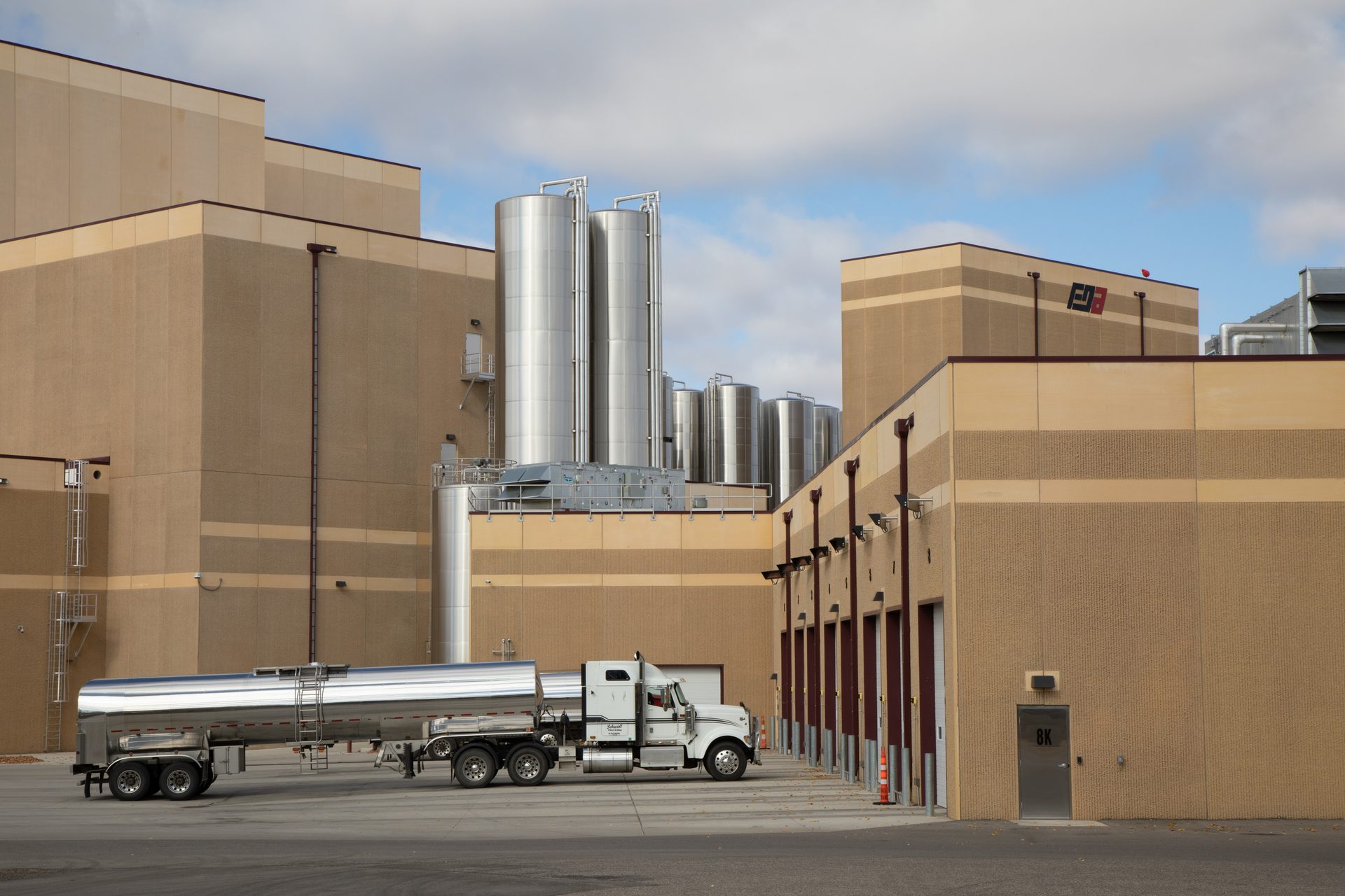 Tan industrial buildings, multiple silver silos, and a semi-truck with a tanker trailer.