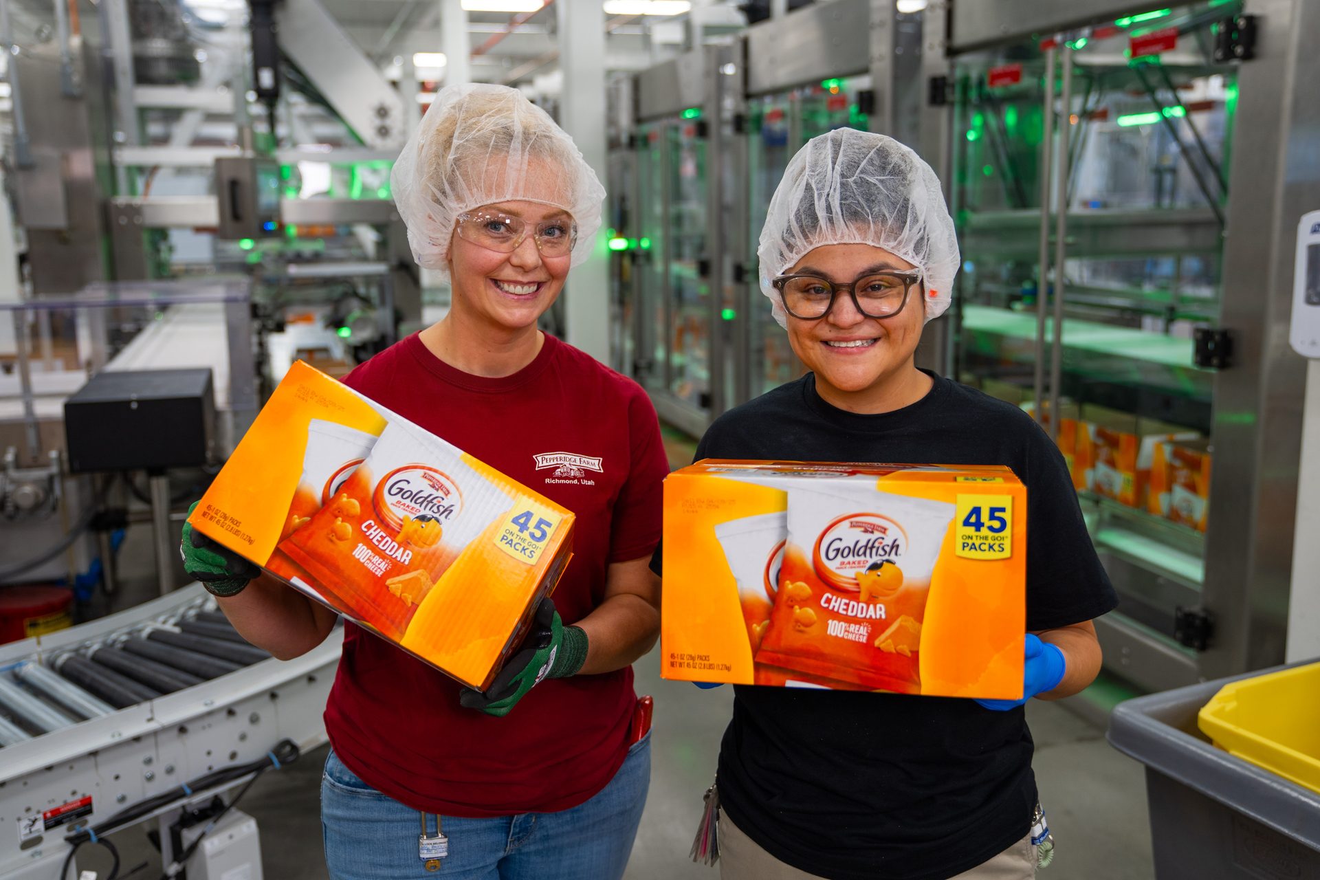 Two smiling women in hairnets hold large boxes of Goldfish crackers in a production facility.