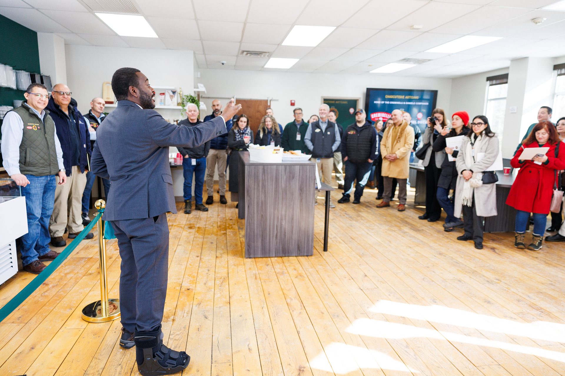 Man in a suit with a medical boot speaks to a diverse group of people indoors at an event.