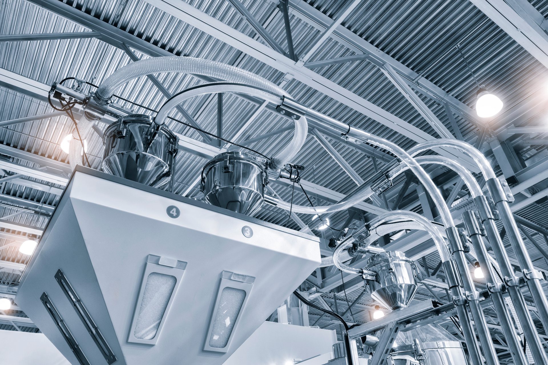 Industrial machinery with hoppers, clear tubes, and a metal ceiling in a modern factory.