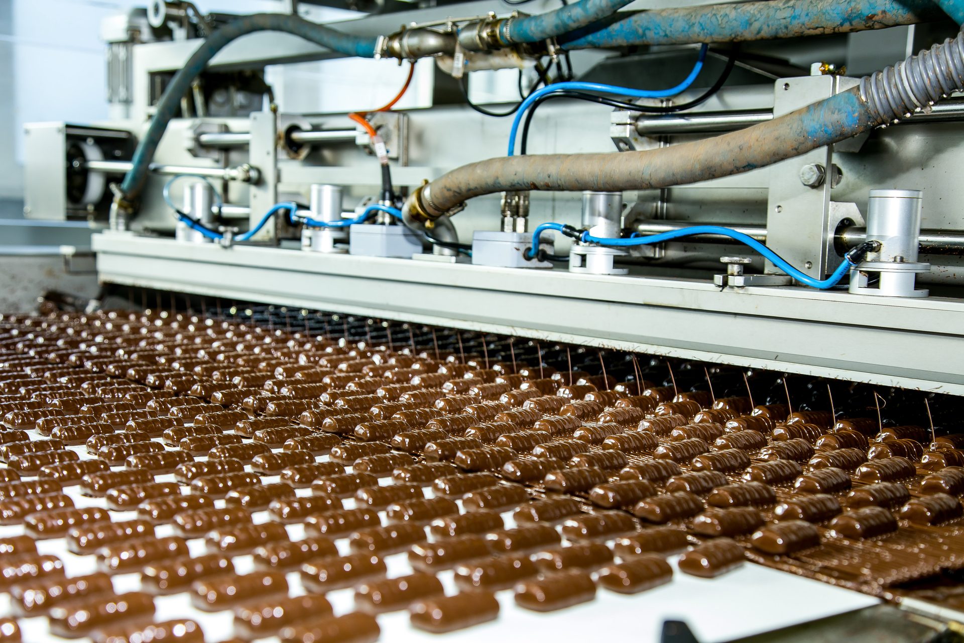 Rows of chocolate candies moving on a conveyor belt, being coated by industrial machinery in a factory.