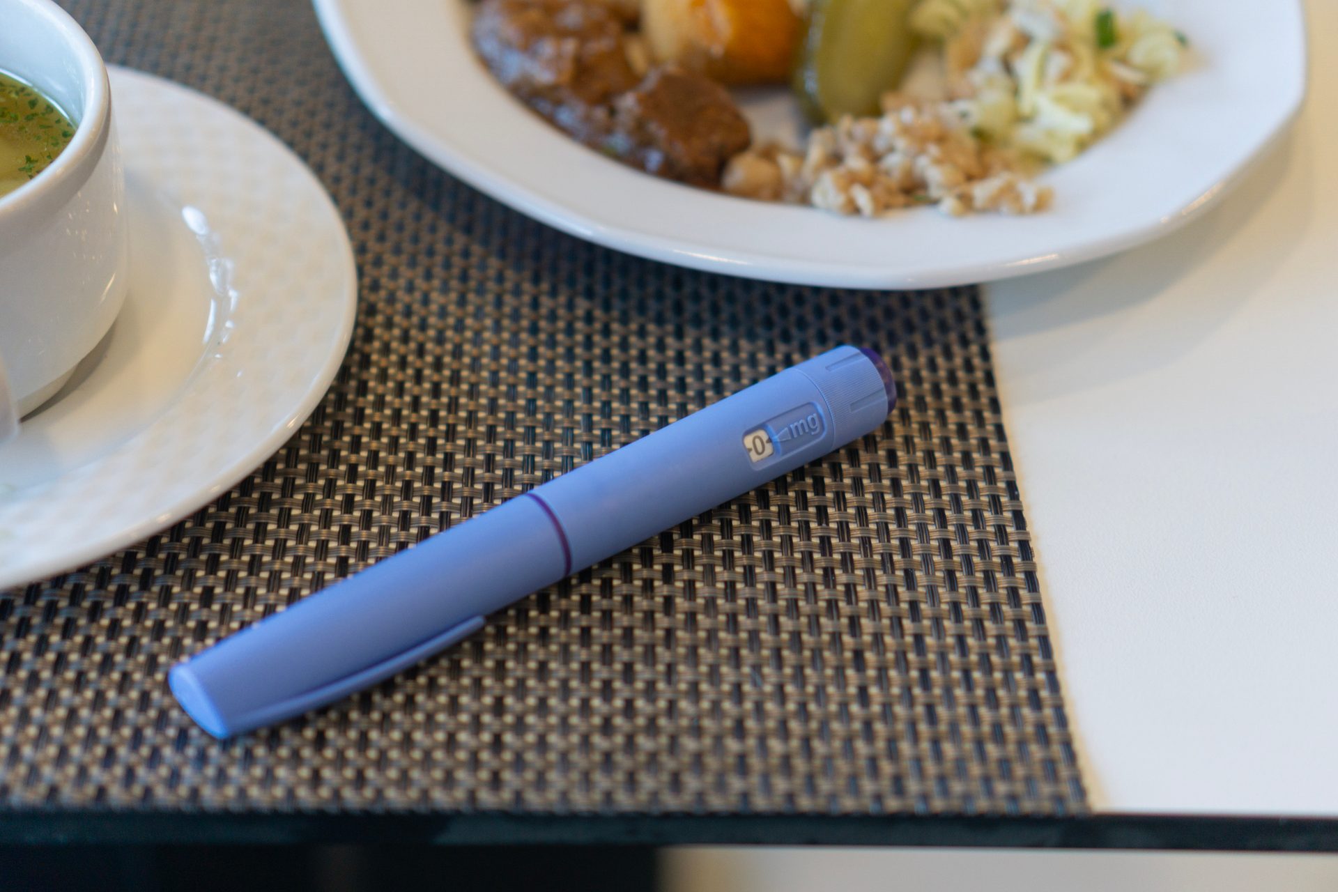 A light purple injection pen is on a placemat next to a blurred cup of soup and a plate of food.