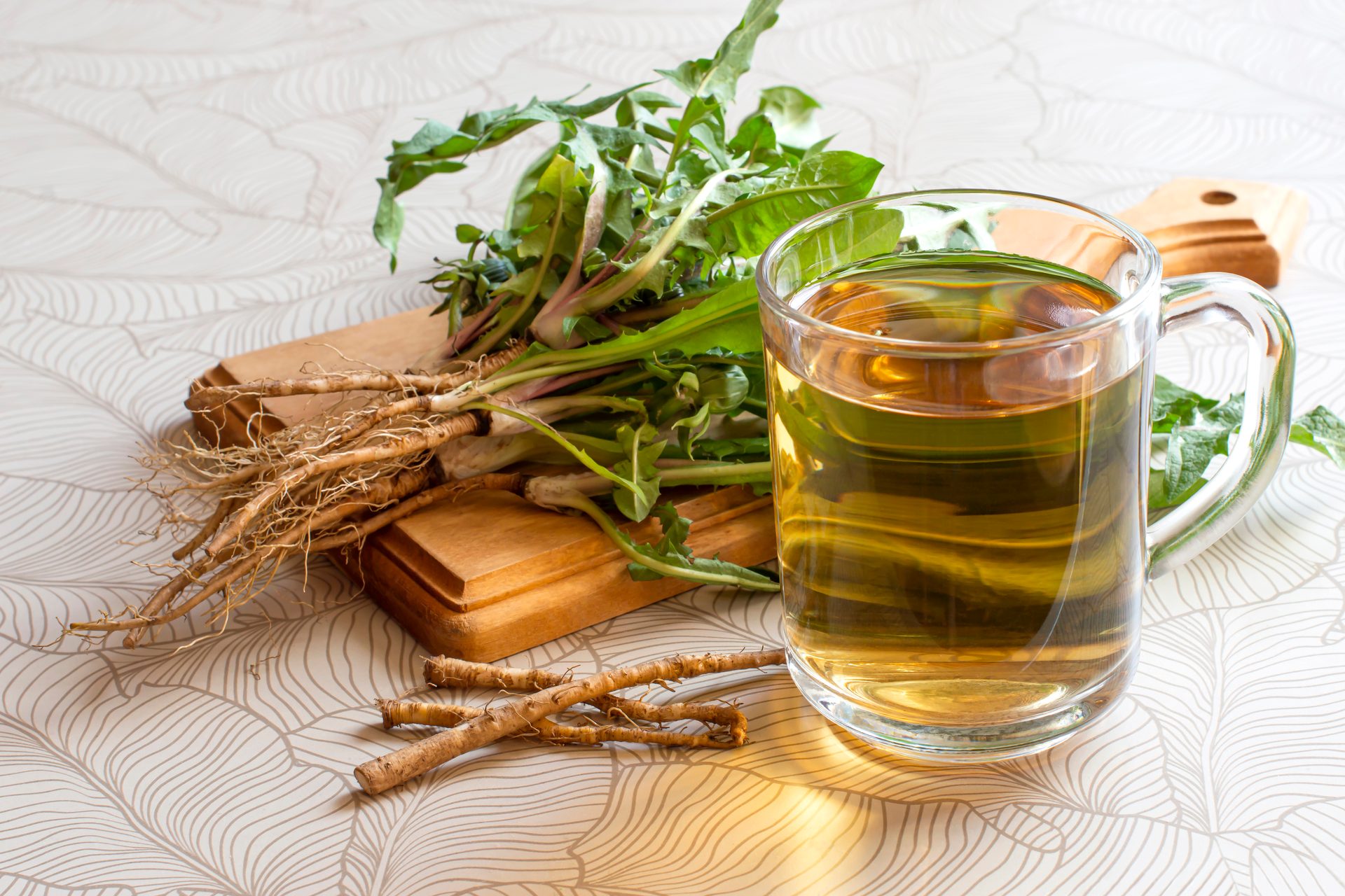 Fresh dandelion roots and leaves next to a clear mug of dandelion tea on a patterned surface.
