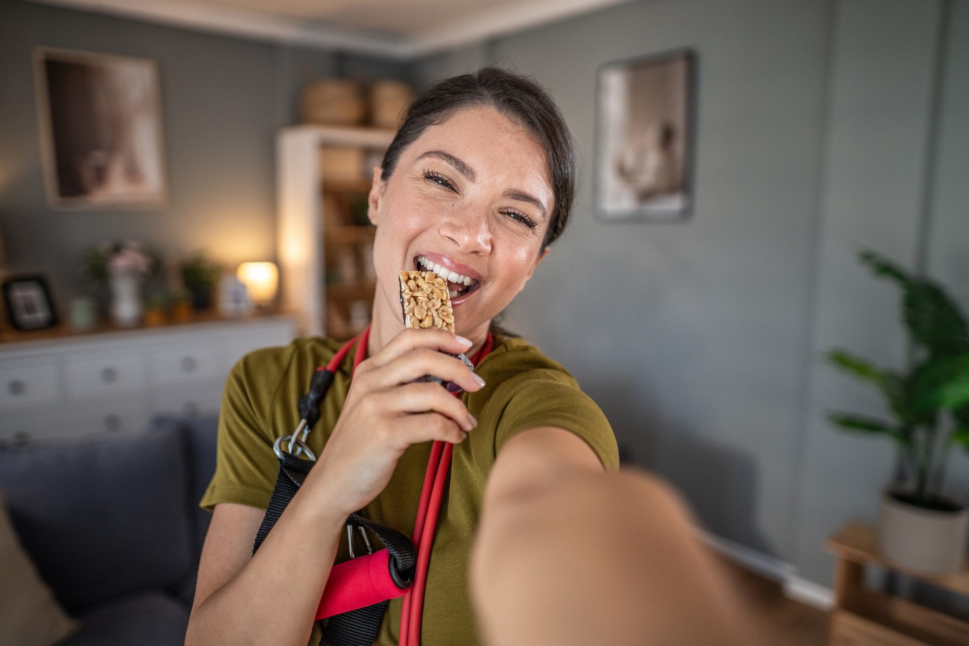 A smiling woman takes a selfie, eating a granola bar with exercise bands around her neck.