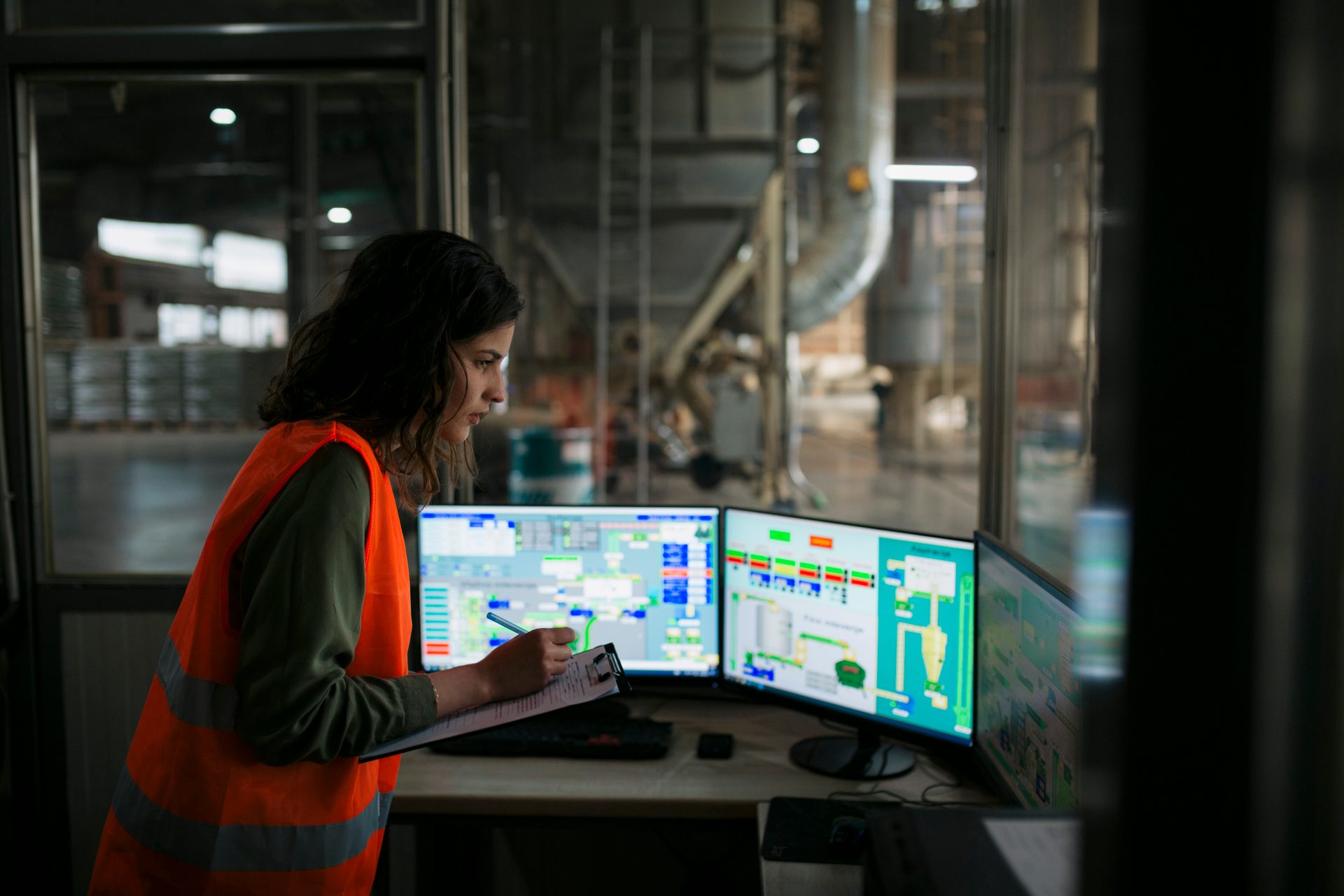 Woman in safety vest monitoring industrial equipment on multiple screens, taking notes.