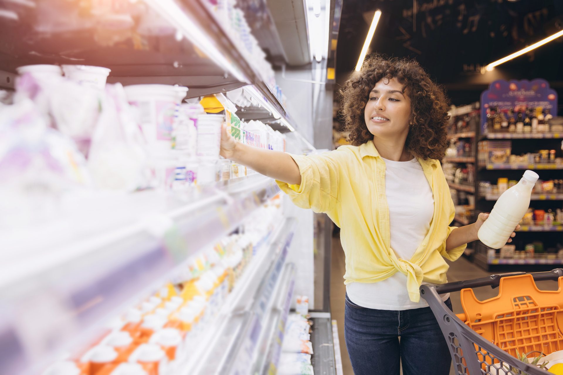 Woman with curly hair shops for dairy products in a supermarket, holding a milk bottle and reaching for yogurt.