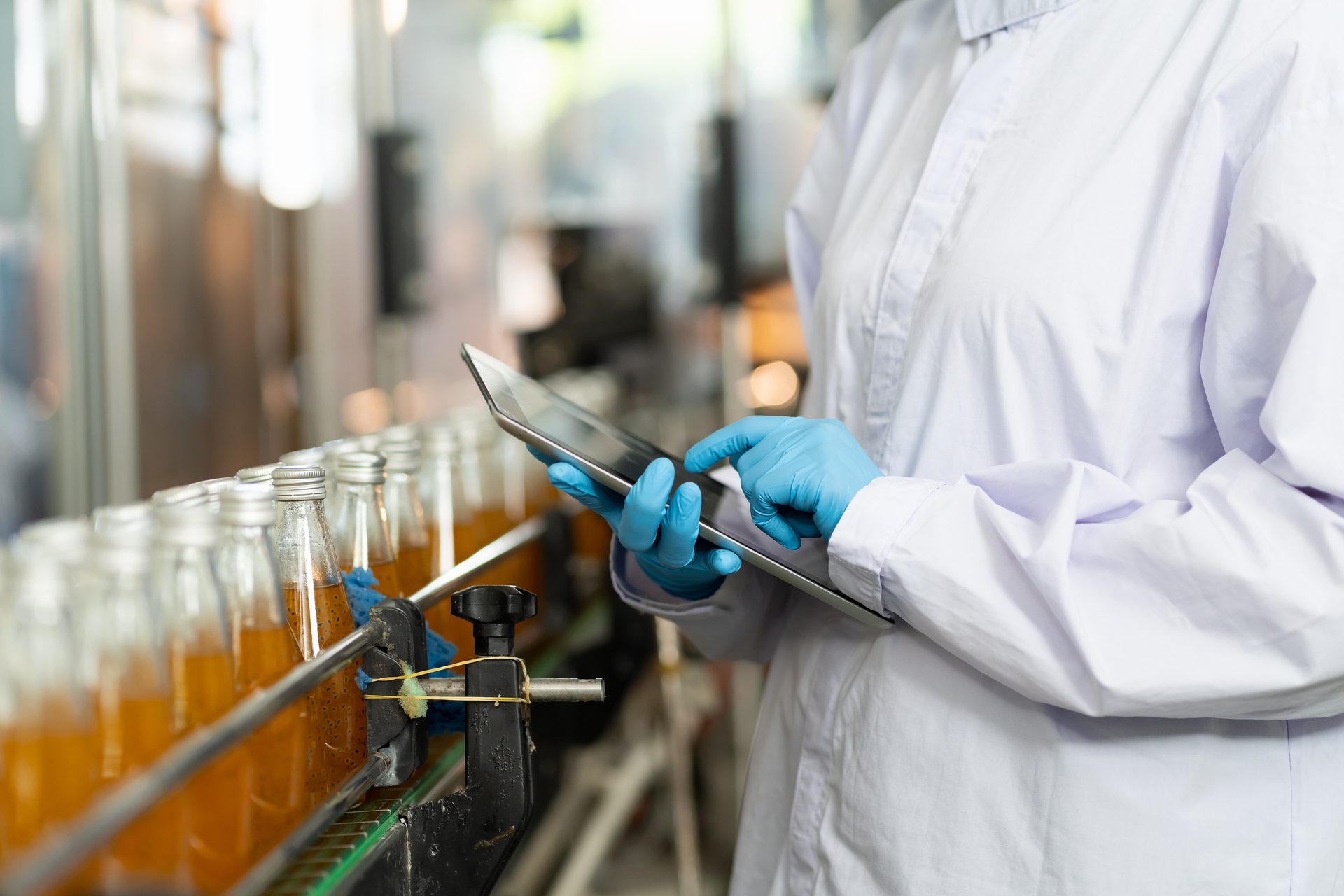 A person in a lab coat and gloves uses a tablet to inspect a beverage production line with bottles.