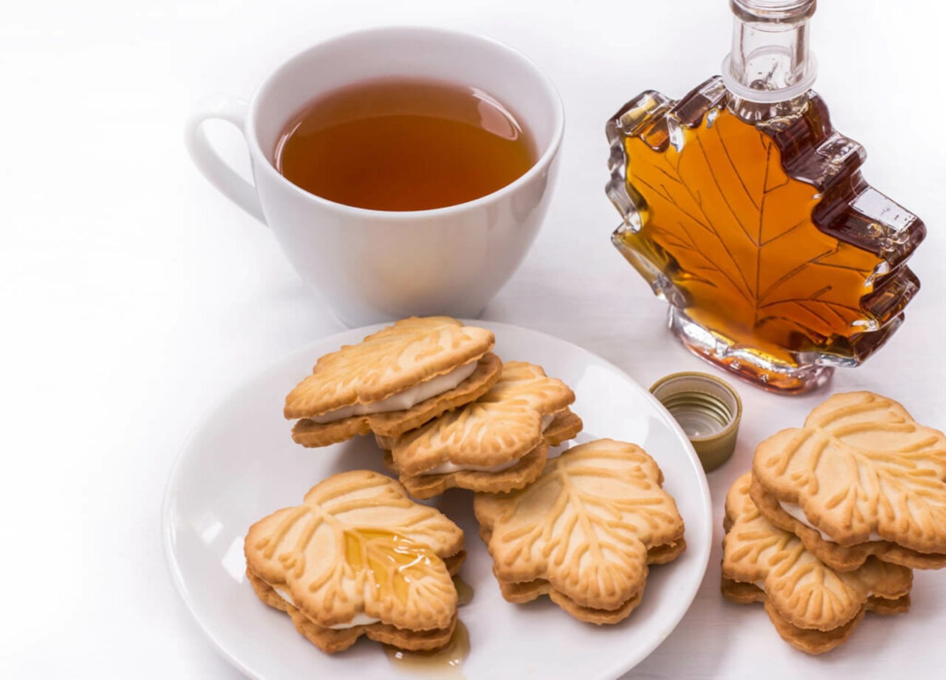 Maple leaf cookies with creamy filling, maple syrup in a leaf-shaped bottle, and a cup of tea.