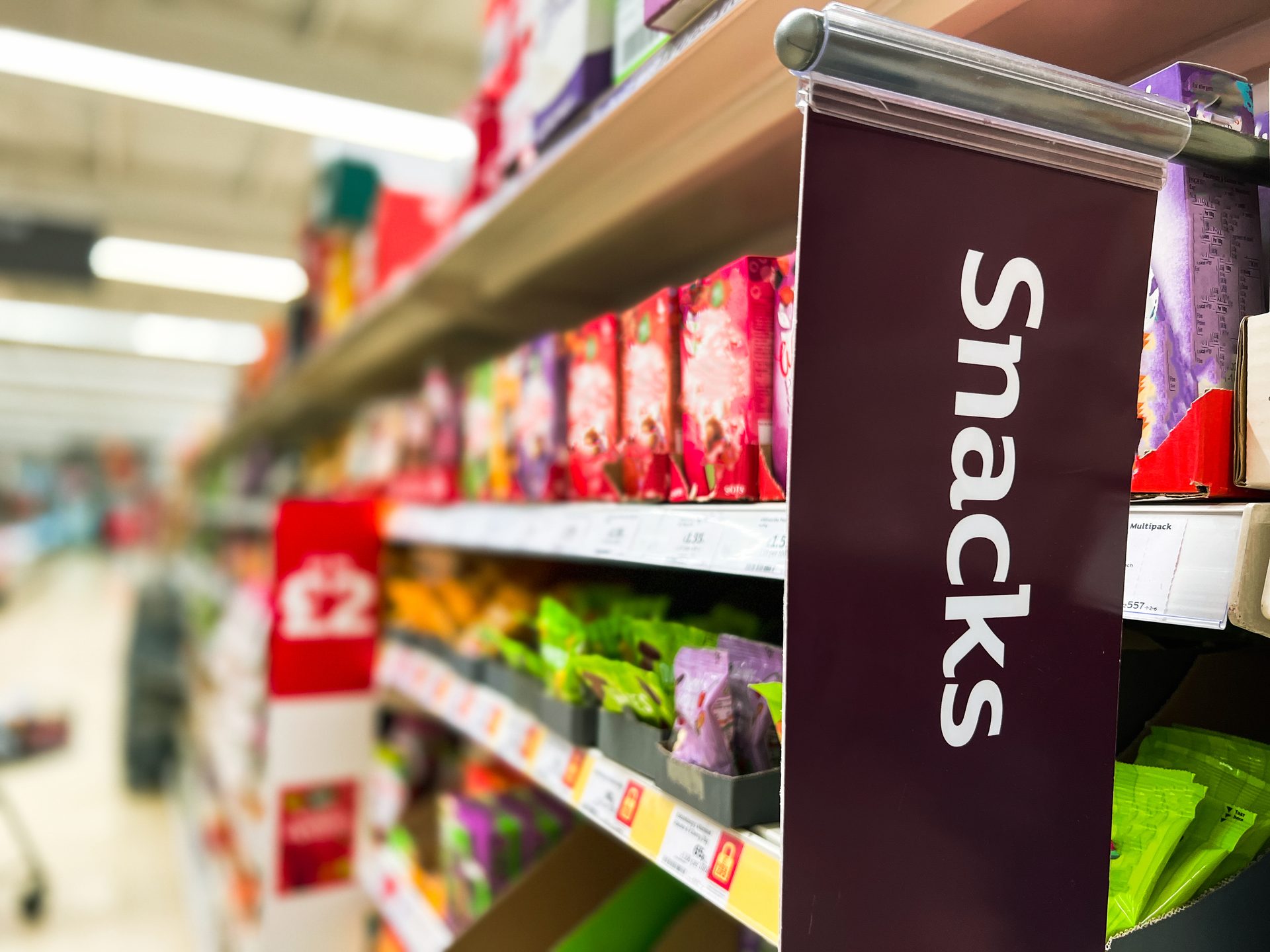 A "Snacks" sign hangs in a supermarket aisle with blurred products on shelves.