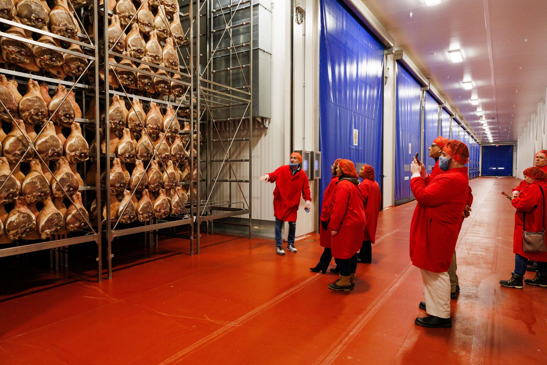 People in red coats tour a large facility with many hams hanging on racks for curing.