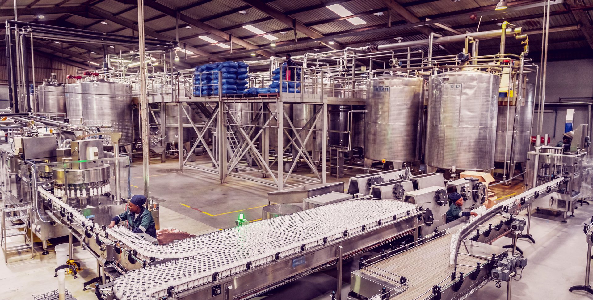 Factory bottling line with workers, tanks, and bottles on conveyors.
