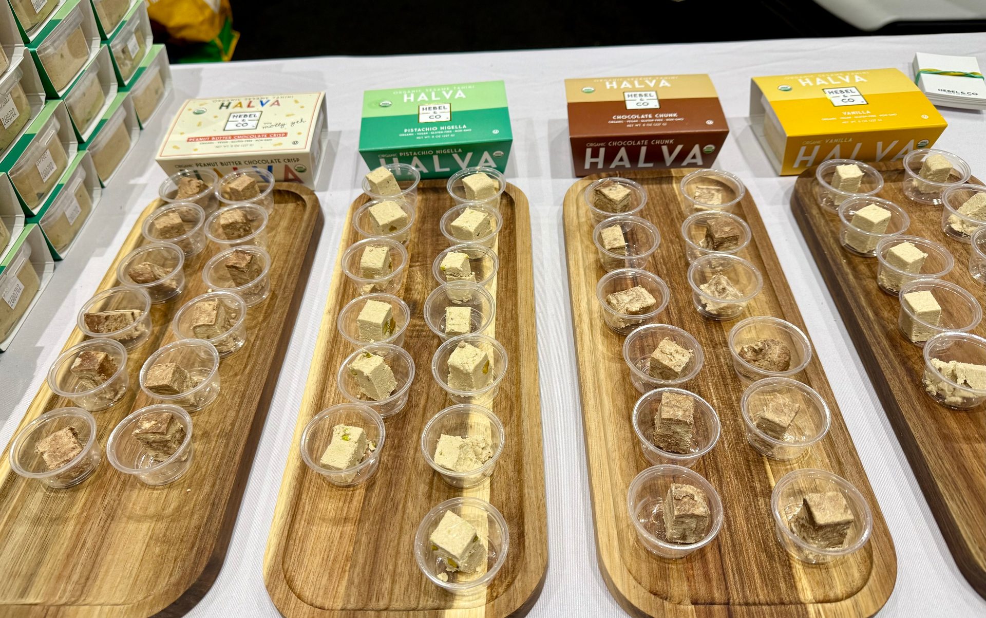 Four wooden trays hold samples of different Halva flavors in small plastic cups, with corresponding product boxes visible behind them.