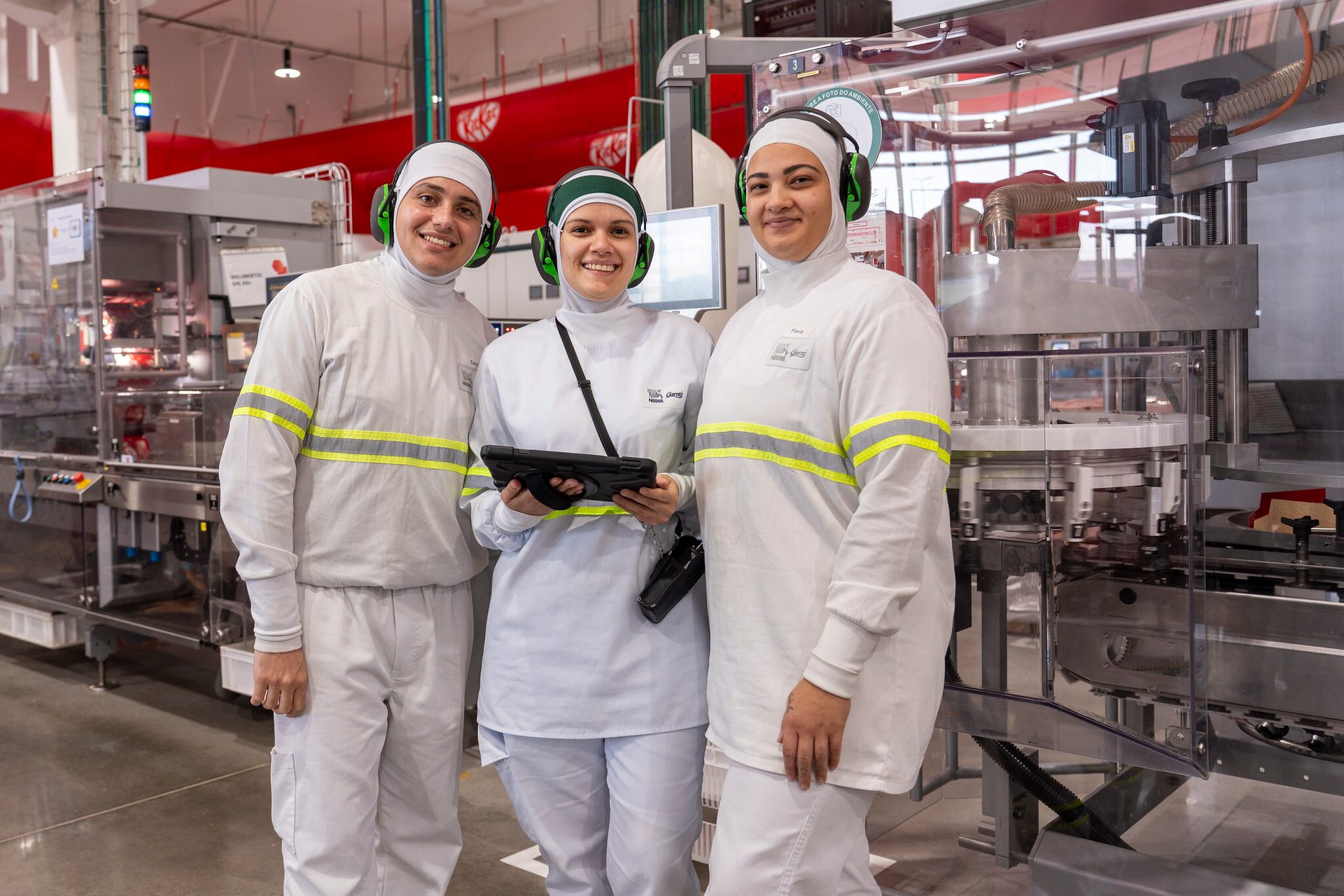 Three smiling KitKat factory workers in uniform with ear protection, one holding a tablet.