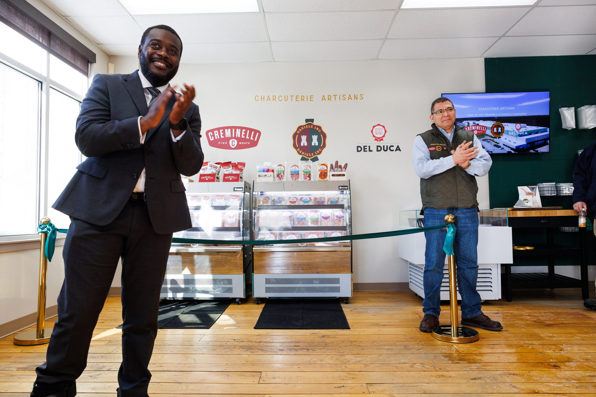 Two men clapping in a store, with charcuterie product displays and brand logos "CREMINELLI" and "DEL DUCA".