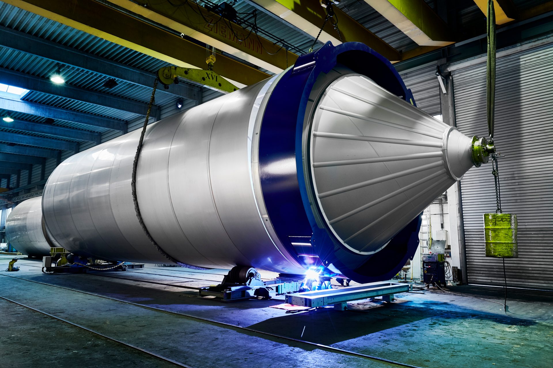 A large metal cylinder, possibly a rocket stage or industrial vessel, undergoing welding in a factory.