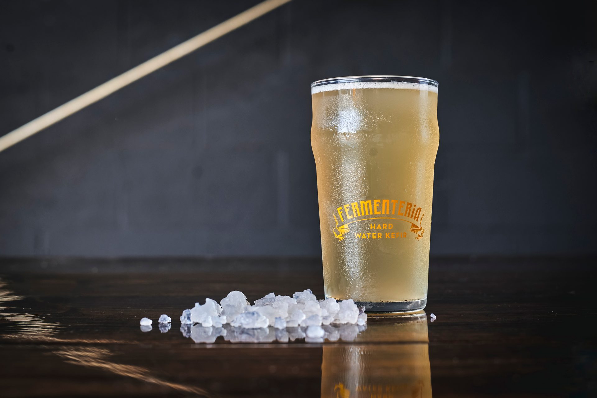 Frosty glass of "Fermenteria Hard Water Kefir" with kefir grains on a dark wood table.