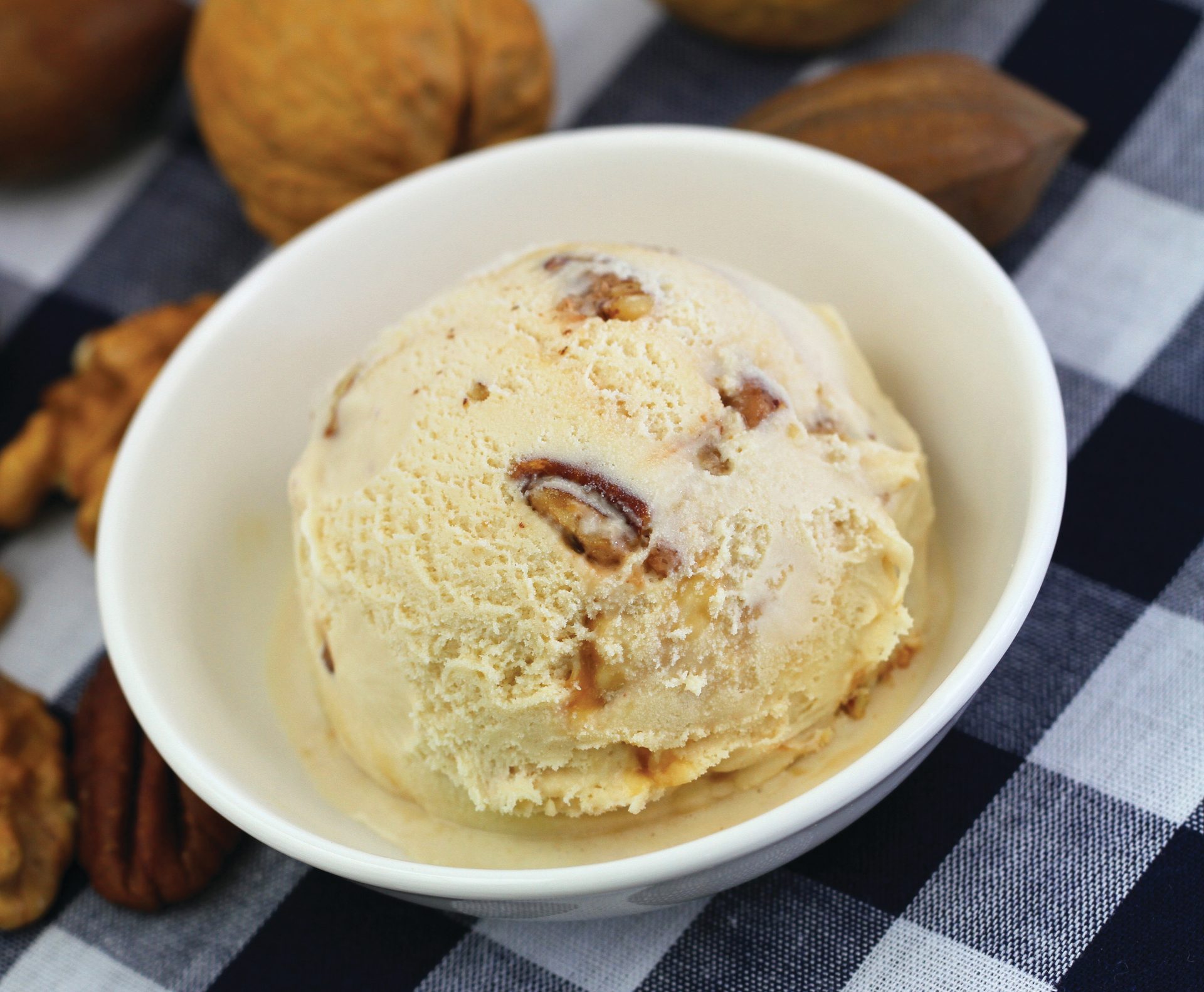 Scoop of pecan ice cream in white bowl on checkered cloth with nuts.