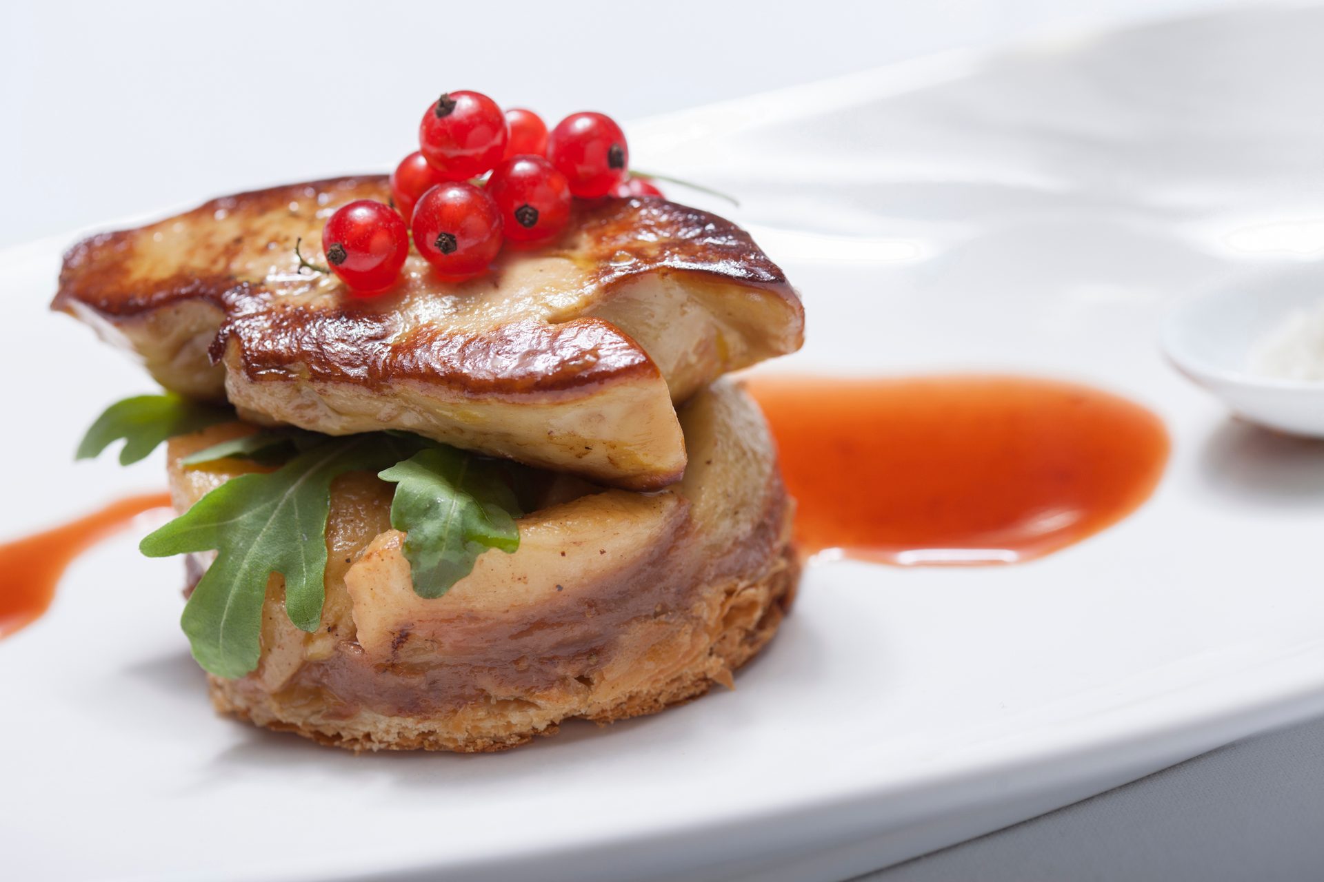 Seared foie gras stack with red currants, arugula, and sauce on white plate.
