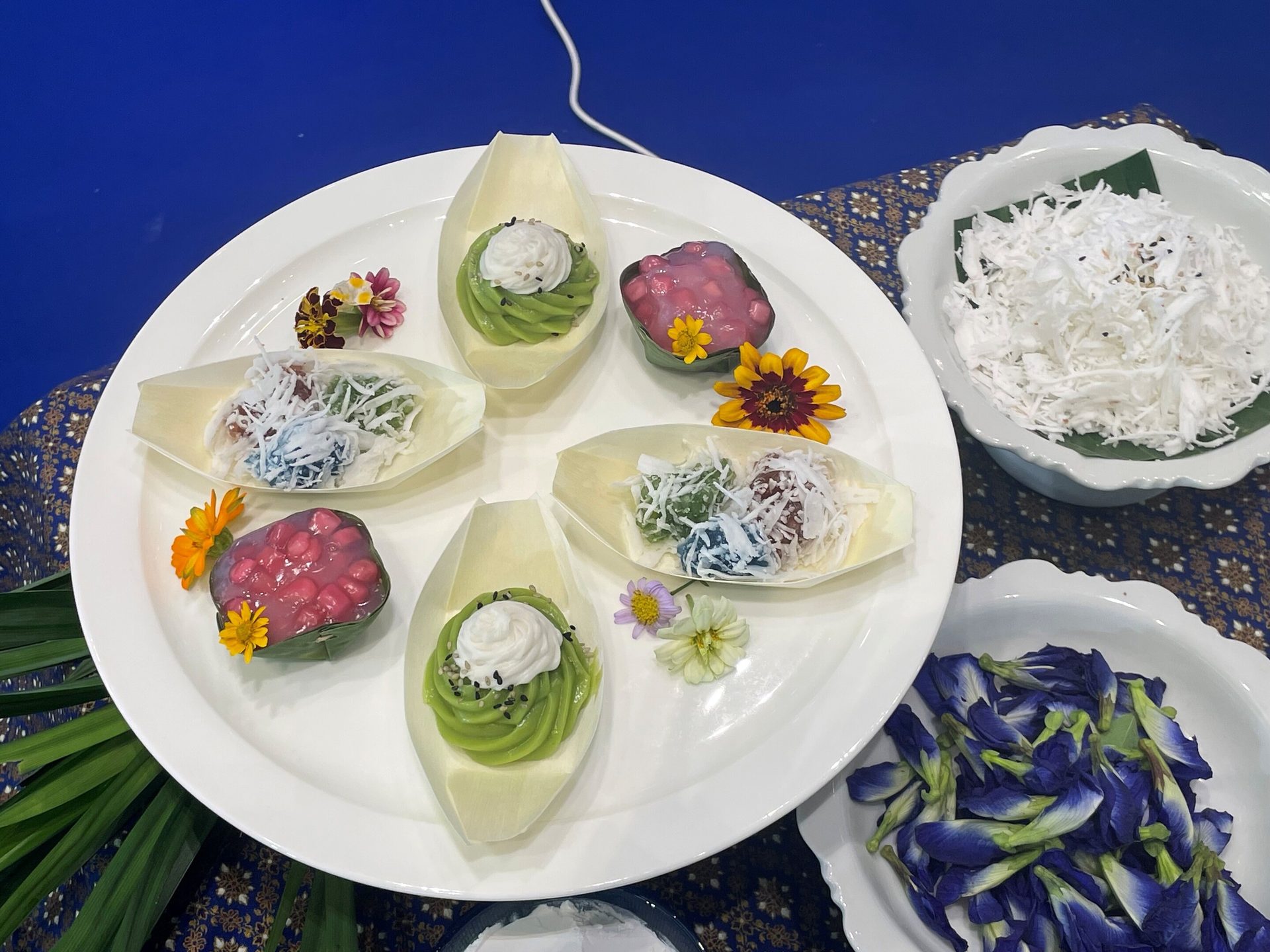 Vibrant Thai desserts, shredded coconut, and butterfly pea flowers artfully arranged on a white platter.