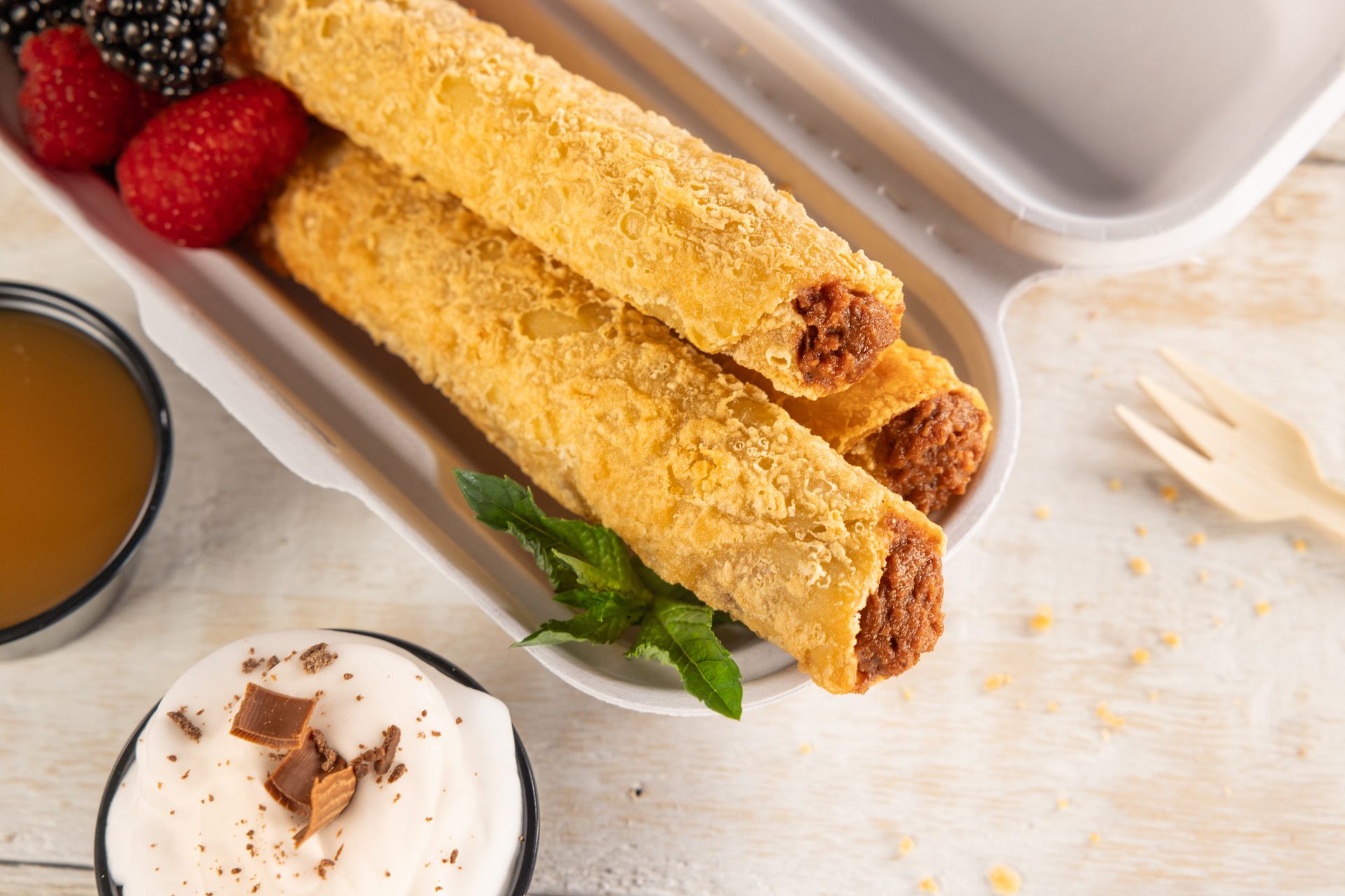 Crispy fried rolls with berries, caramel sauce, and whipped cream in a takeout box.