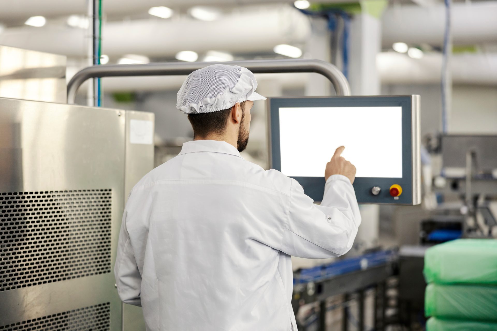 Person in a lab coat and hairnet operating a touchscreen in a factory.