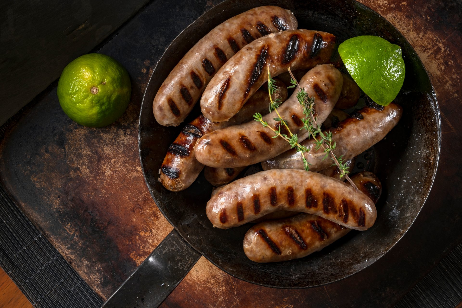A cast-iron pan filled with grilled sausages and thyme, alongside a lime, on a rustic wooden board.