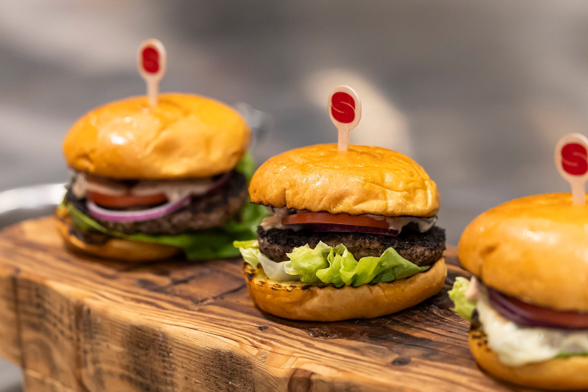 Three mini burgers with picks, lettuce, tomato, and onion, on a wooden board.