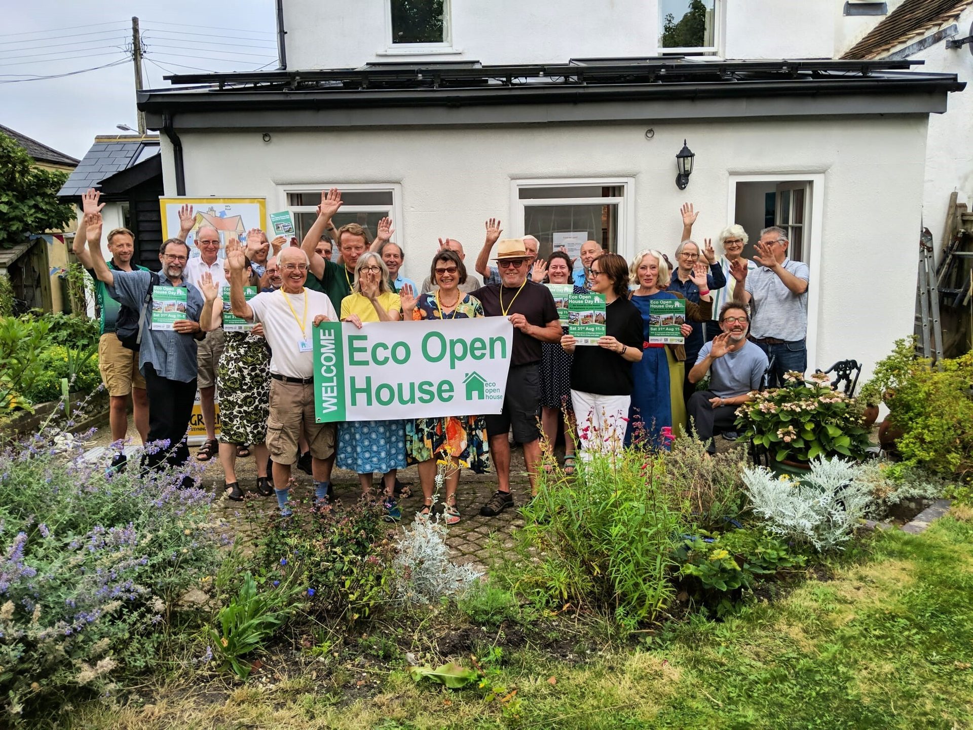 Group waves with "Eco Open House" banner in front of a house and garden.