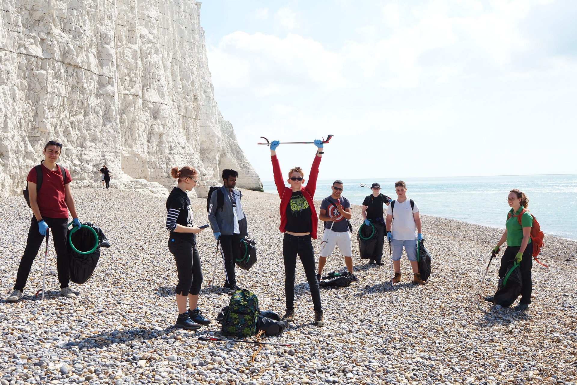 Group doing a beach cleanup with grabbers and bags on a pebbly beach below white cliffs.