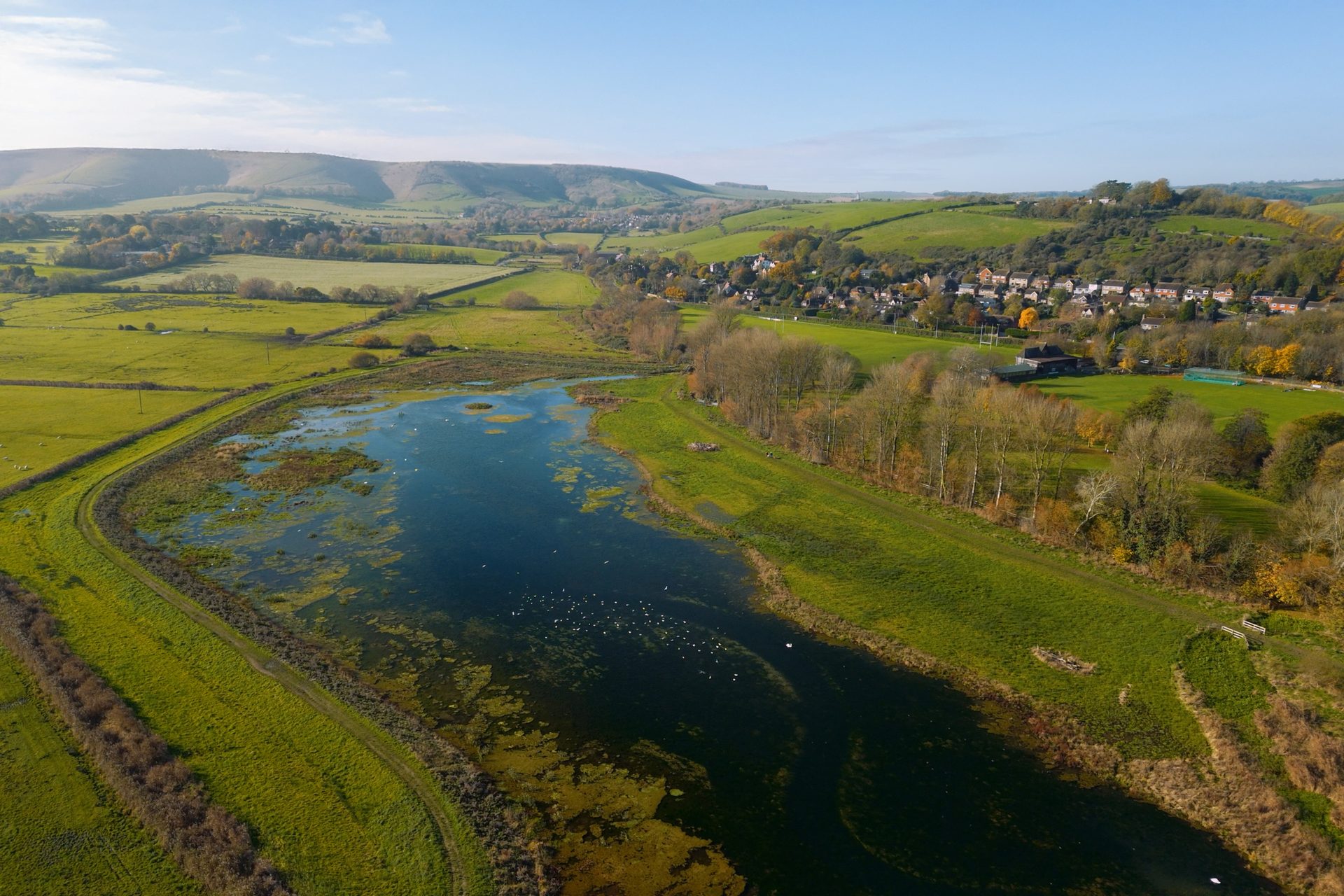 Aerial: green landscape with a lake, rolling hills, and a village, bathed in sunlight.