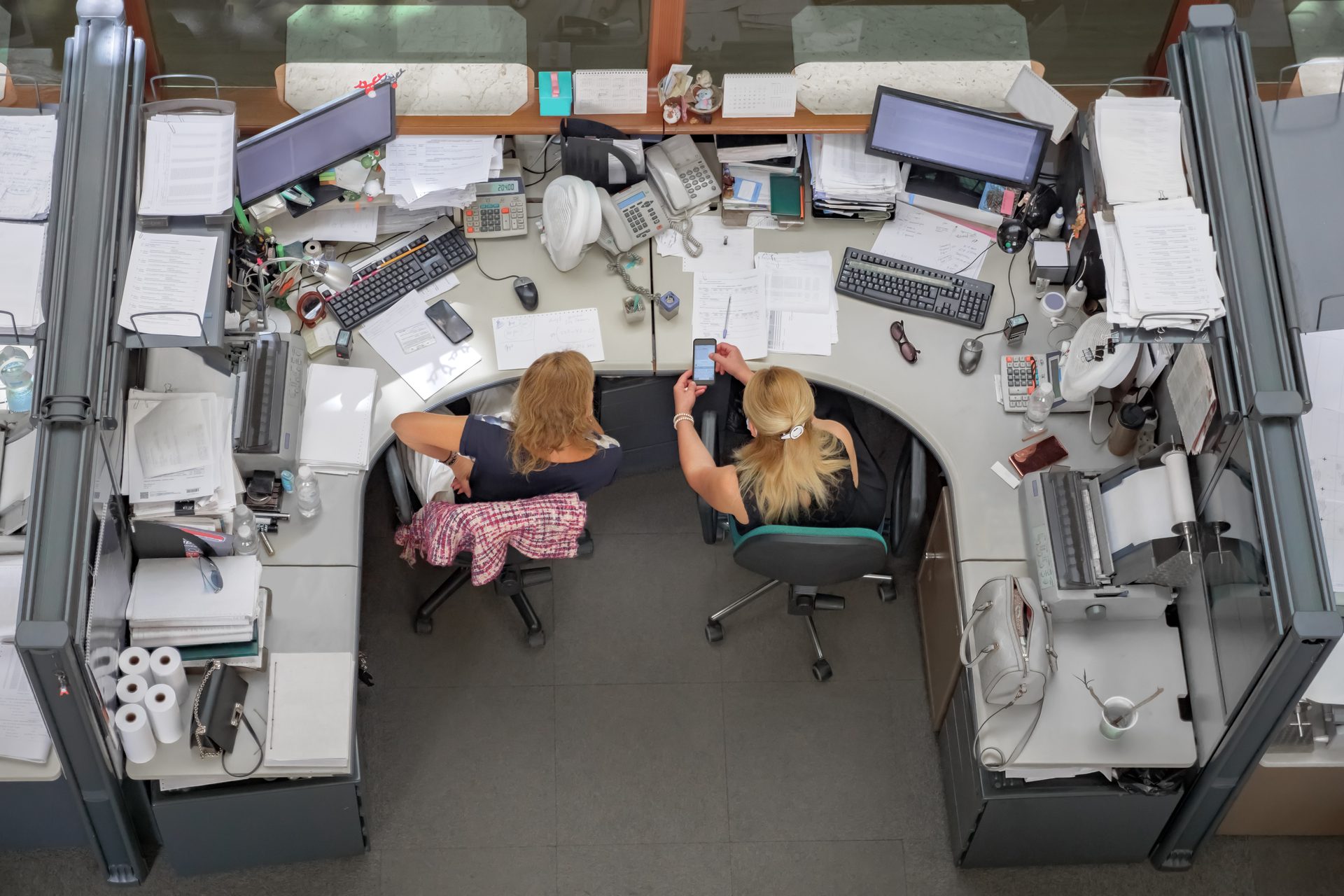 Overhead view of two women at a cluttered office desk with computers, phones, and papers.