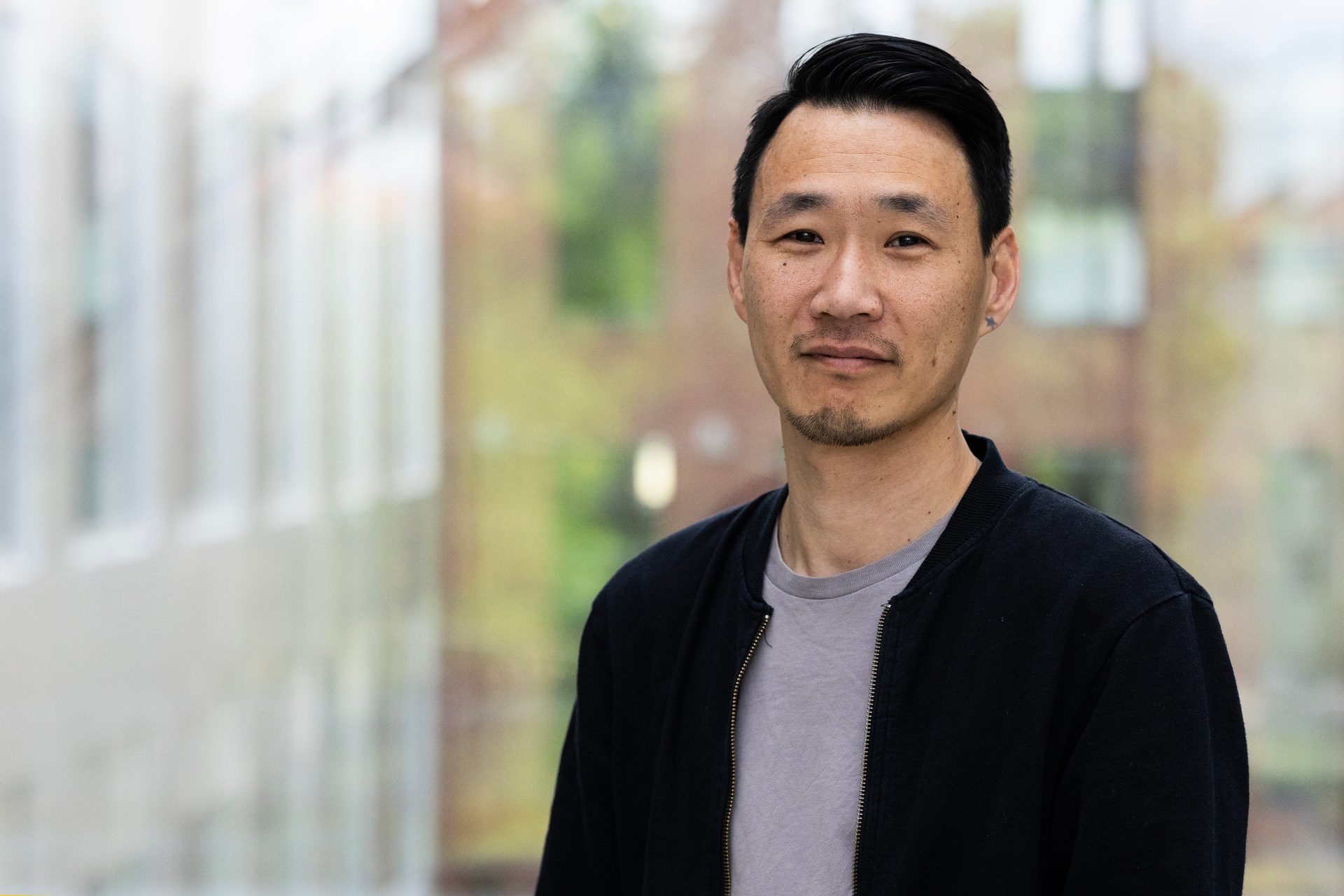 Headshot of an Asian man with short dark hair, wearing a black jacket over a grey t-shirt, smiling slightly.