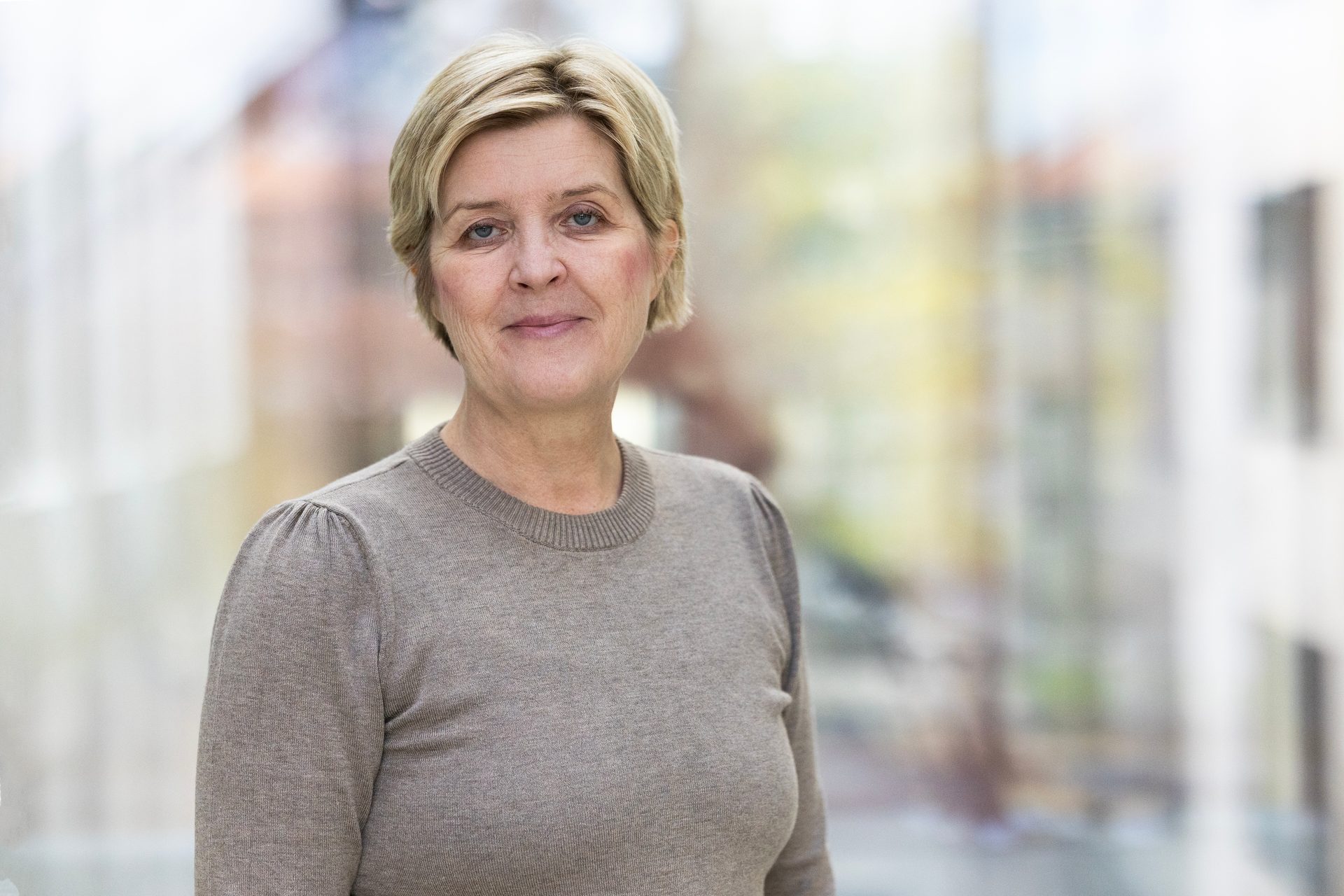 A smiling middle-aged woman with short blonde hair in a brown sweater against a blurred background.