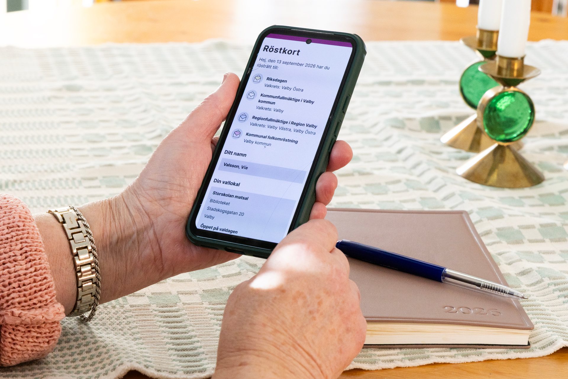Person holding a smartphone displaying election card information next to a pen and planner.