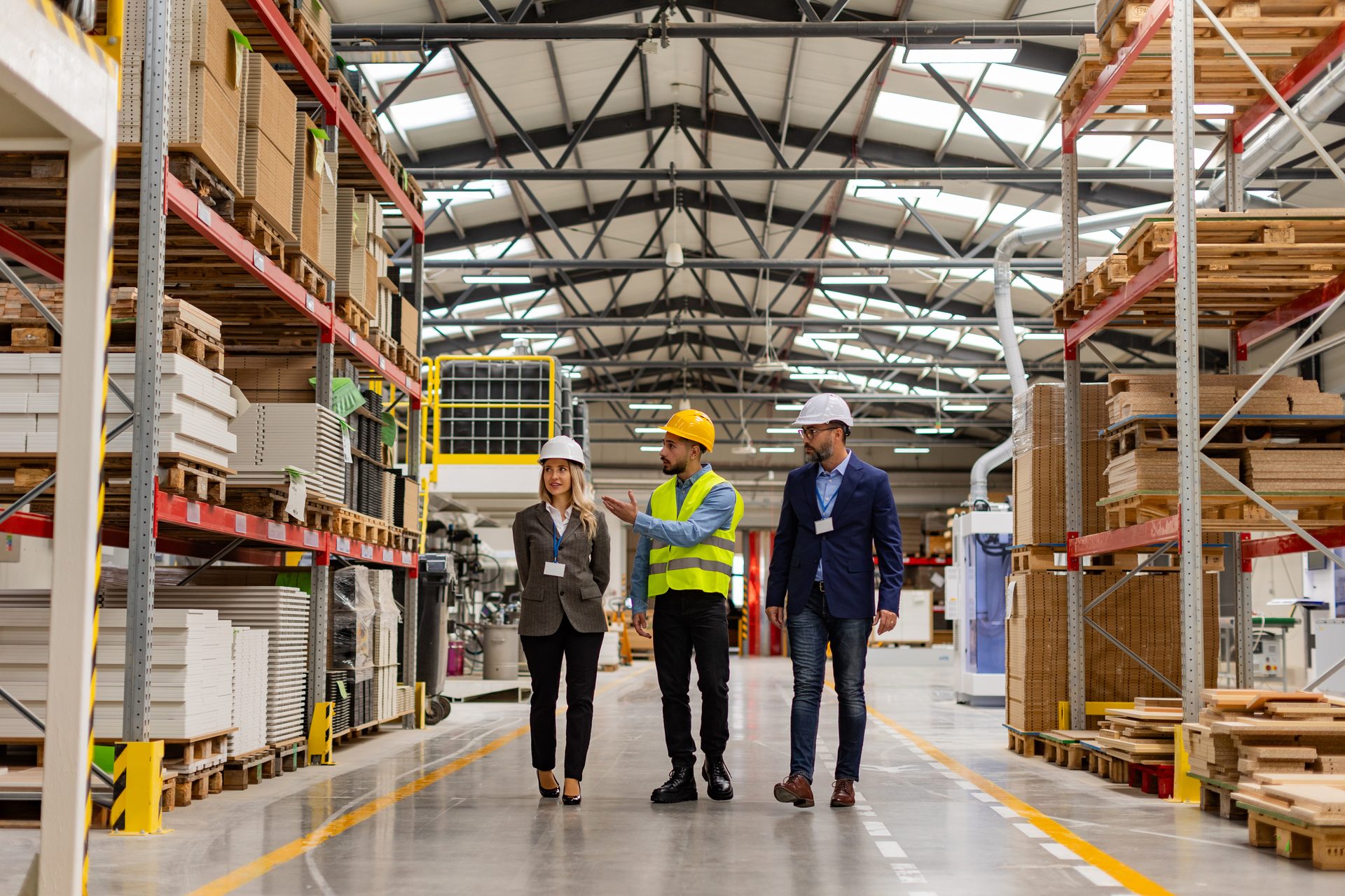 Three professionals, including a supervisor in a hi-vis vest, discussing in a large industrial warehouse.