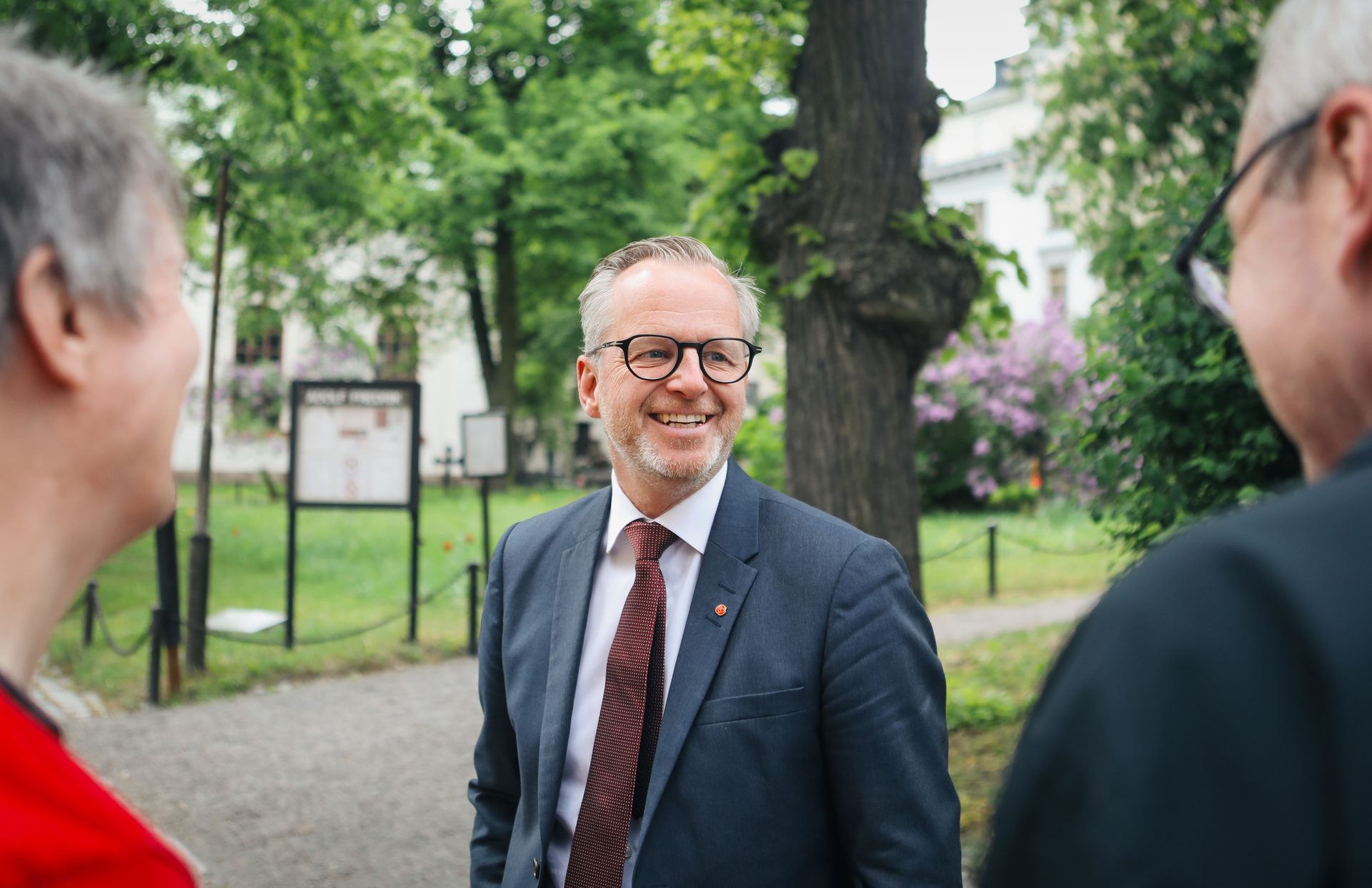 A man in a suit and glasses smiles, conversing with two people in a park setting.