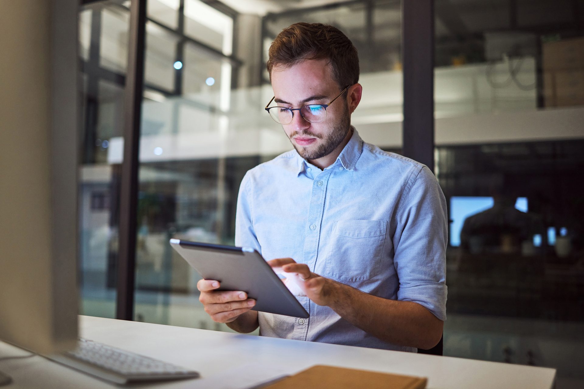 A young man in glasses is intently working on a tablet at a desk in a modern office environment.
